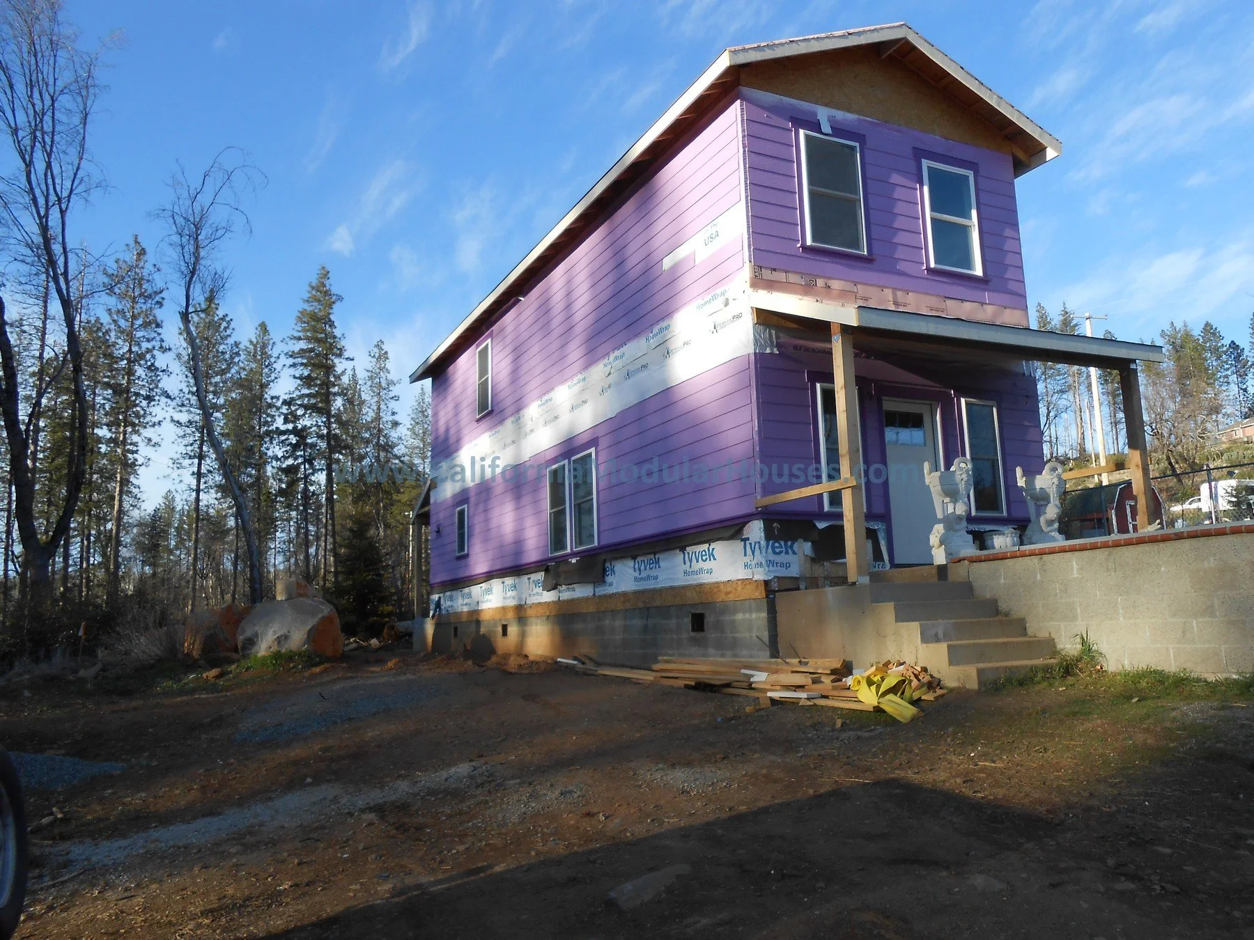 A two-story house under construction with purple siding, a porch with two chairs, and surrounded by trees on a clear day.  Cobb Mountain, Lake County, California.  Modular Homes in California, Modular Homes California,