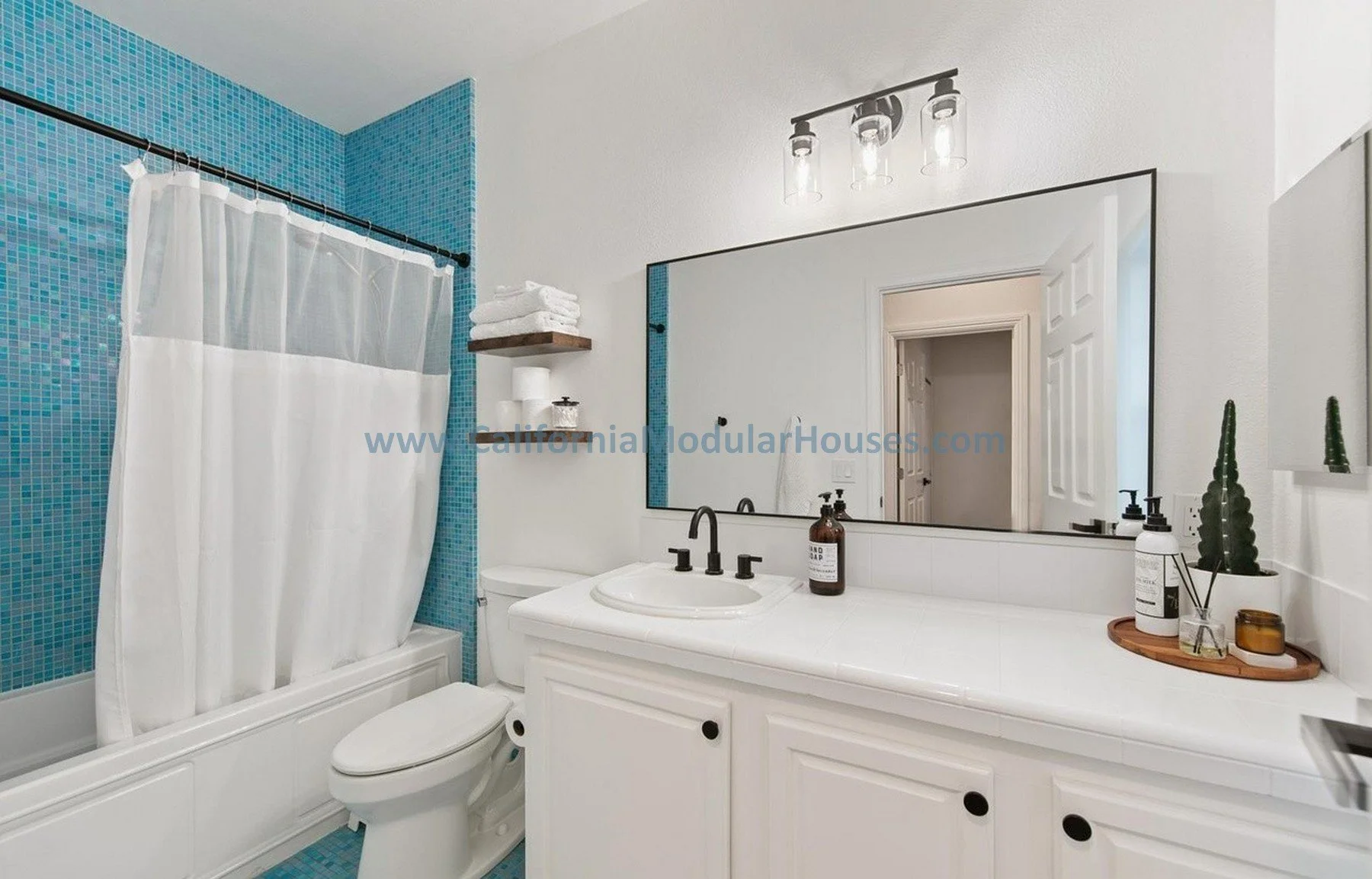 A modern bathroom with white vanity, large mirror, and black fixtures. Blue mosaic tile shower with a white curtain. Decorated with potted plants and toiletries.