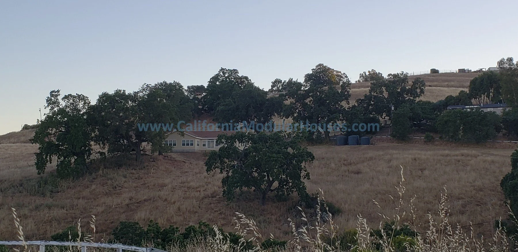 A hillside with dry grass, trees, and a house with a red roof near the center. The house has multiple windows. A few large trash bins are on the right side of the house. Behind the house, more trees and structures are visible. Modular homes.  