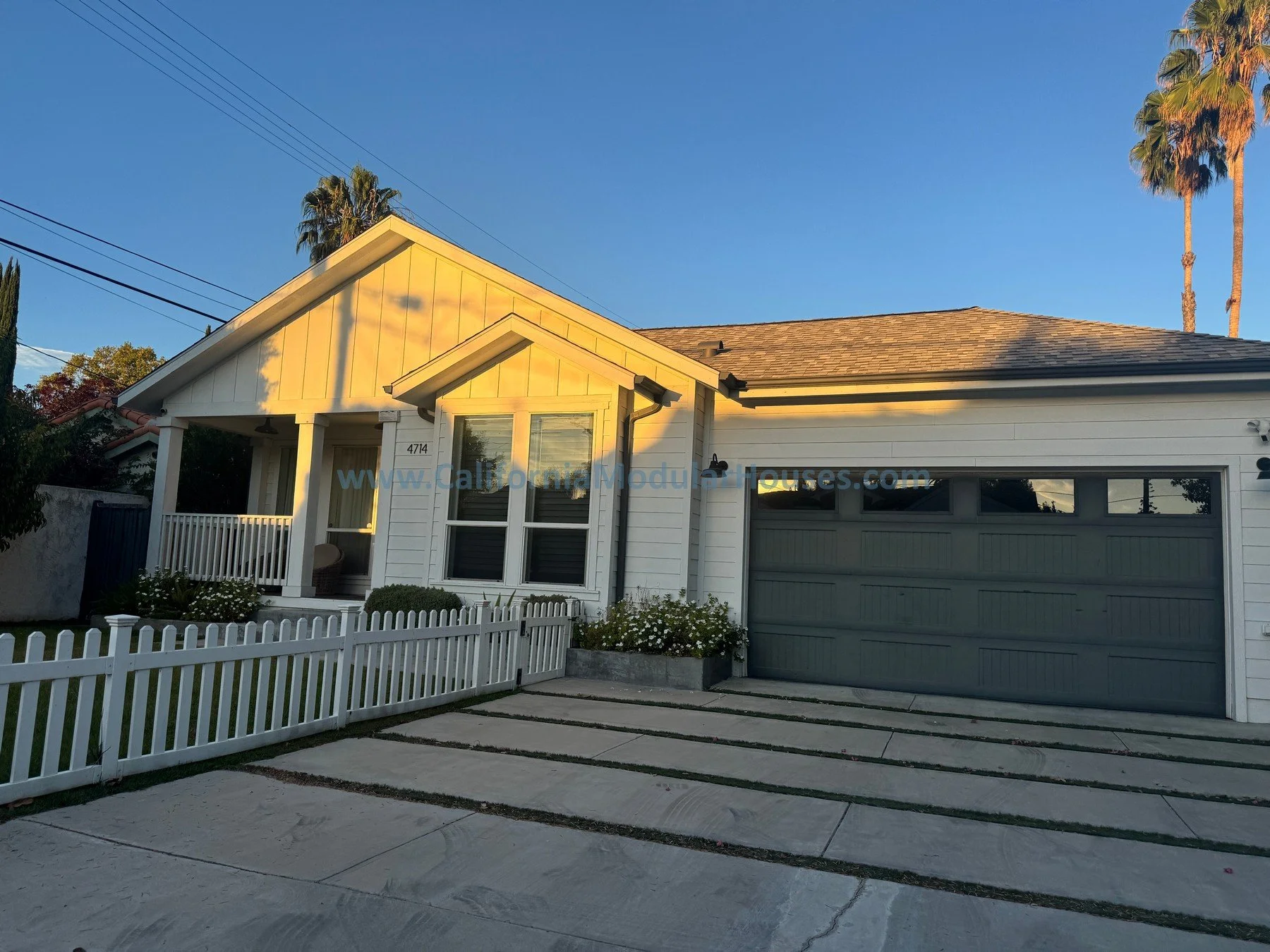 Front view of a modern white house with a gray garage door, a small porch, a white picket fence, and tall palm trees against a clear blue sky during sunset.