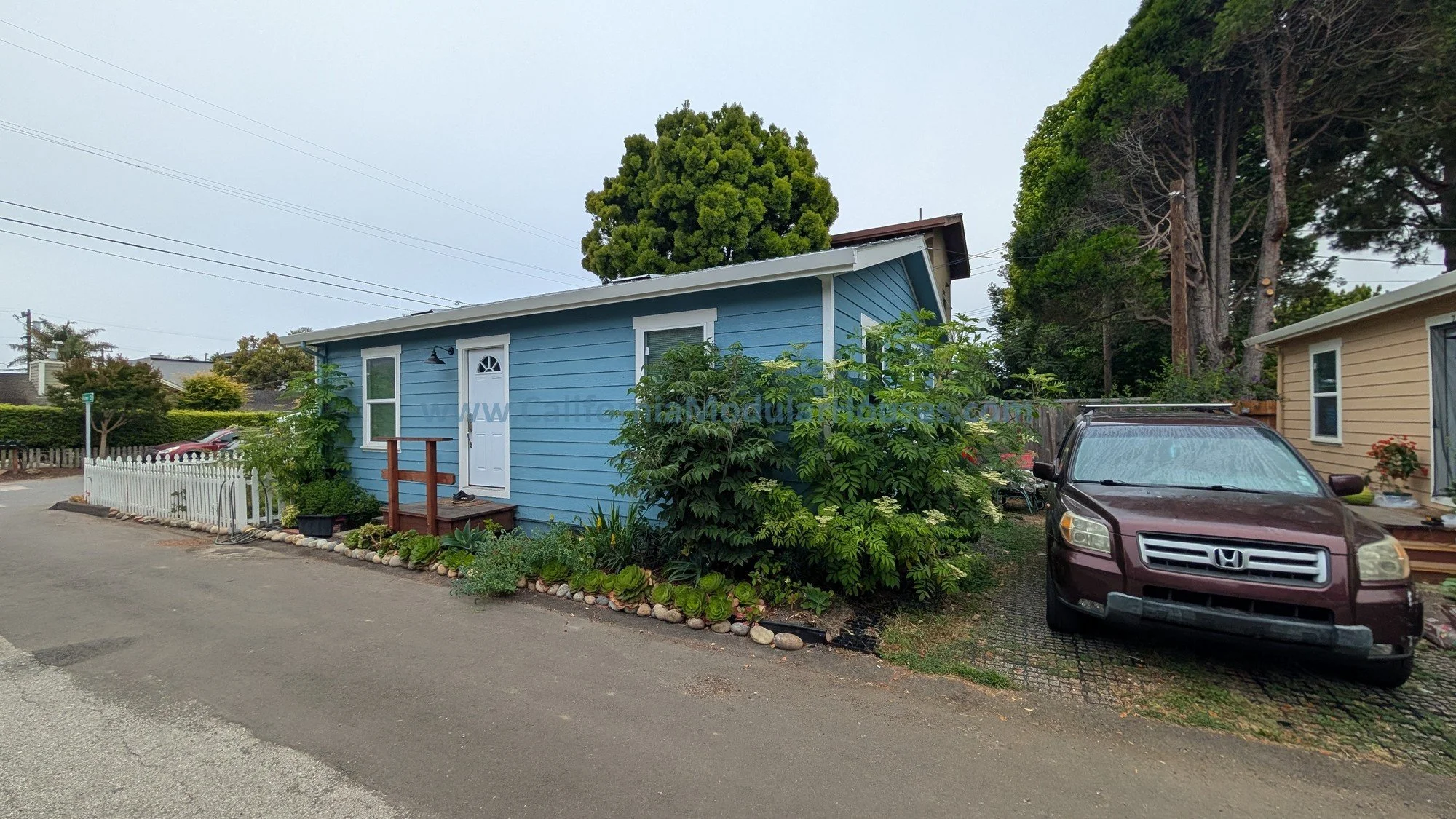 A small blue house with white trim, a white door with a small window, and gray roof. A small wooden ramp leads to the door. There are plants along the house with a white picket fence on the left. On the right, there is a brown Honda SUV parked on a b