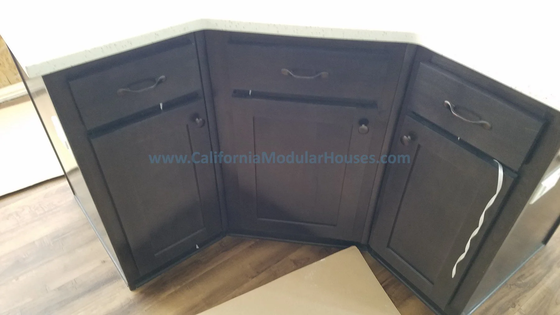 Dark wood kitchen cabinets with black handles and a label 'www.CaliforniaModularHouses.com' printed across the middle, built onto a curved wall section in a home kitchen.  Modular Home CA, California Modular.