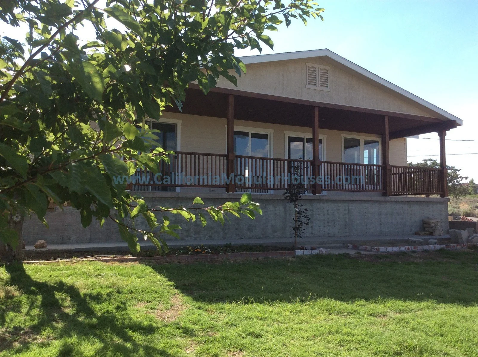 A house with a large front porch, wood railing, beige siding, and white-framed windows, set against a green lawn and trees under a clear blue sky.
