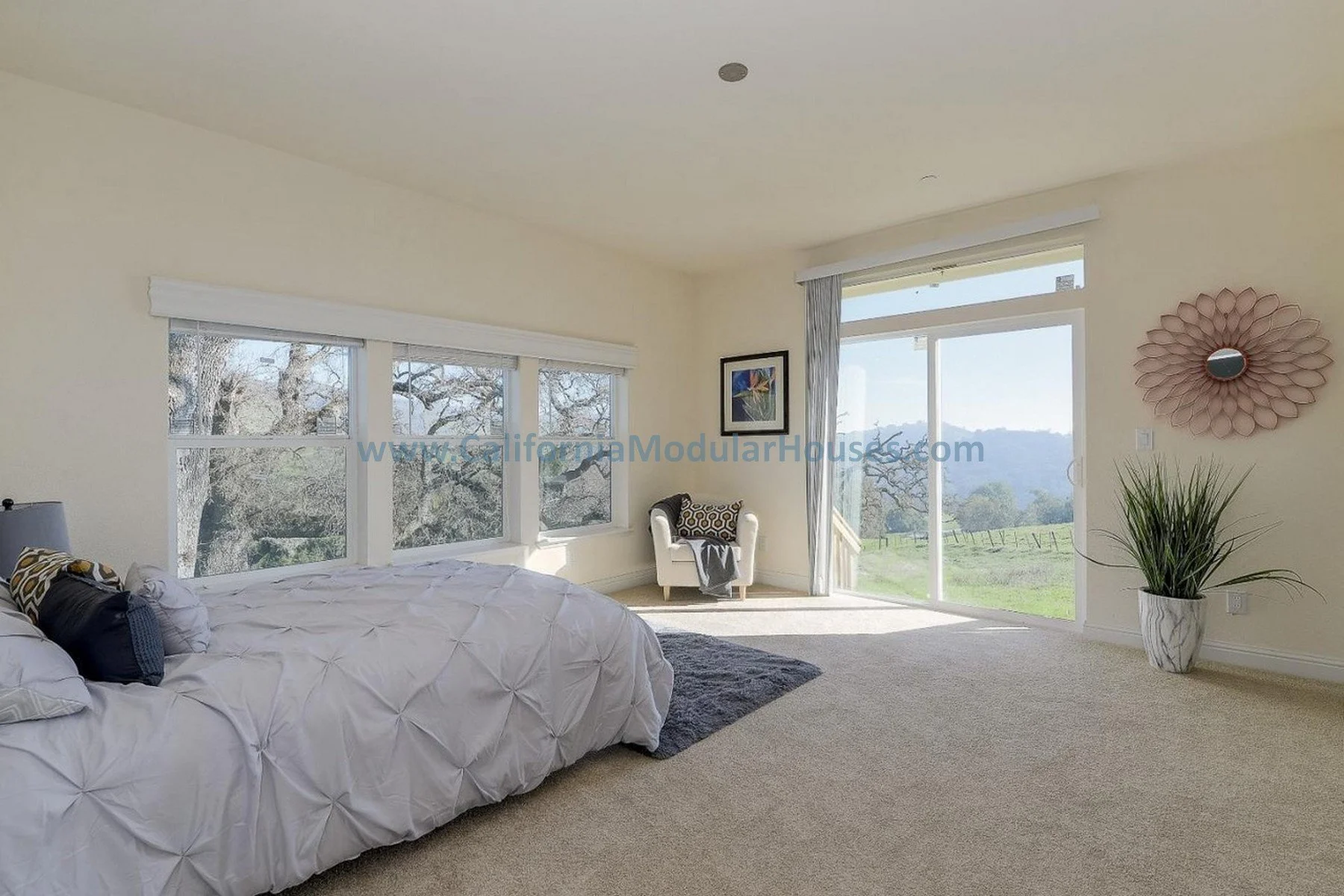 Bright bedroom with large windows overlooking trees and hills, white bed with decorative pillows, armchair, framed artwork, potted plant, and a decorative wall mirror.  Modular Homes, Modular Homes California.