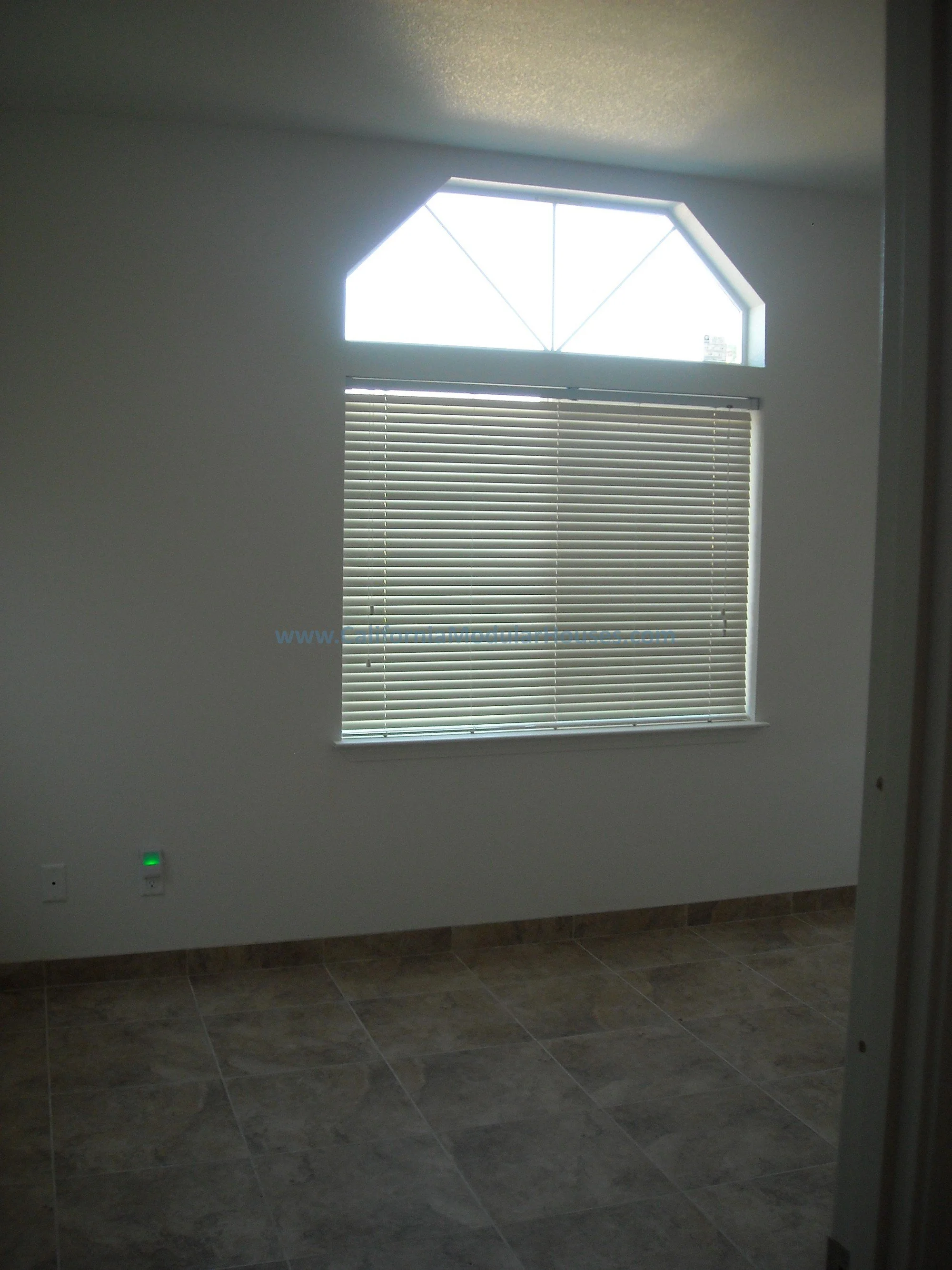 Empty room with a window featuring blinds and a geometric upper window, tiled floor, and electrical outlets on the wall.