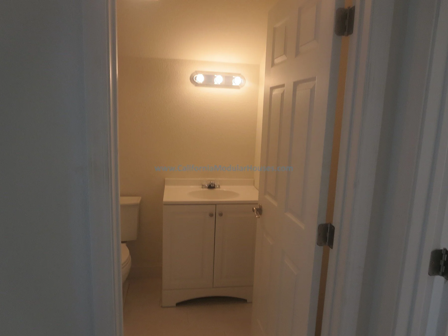 A small bathroom with a white vanity, a mirror, and a light fixture above. Part of the toilet is visible on the left side of the image. The door to the bathroom is open.  Pre-fab Modular.  Cape Cod.  Modulars for Napa.  Napa County, CA. Modular.