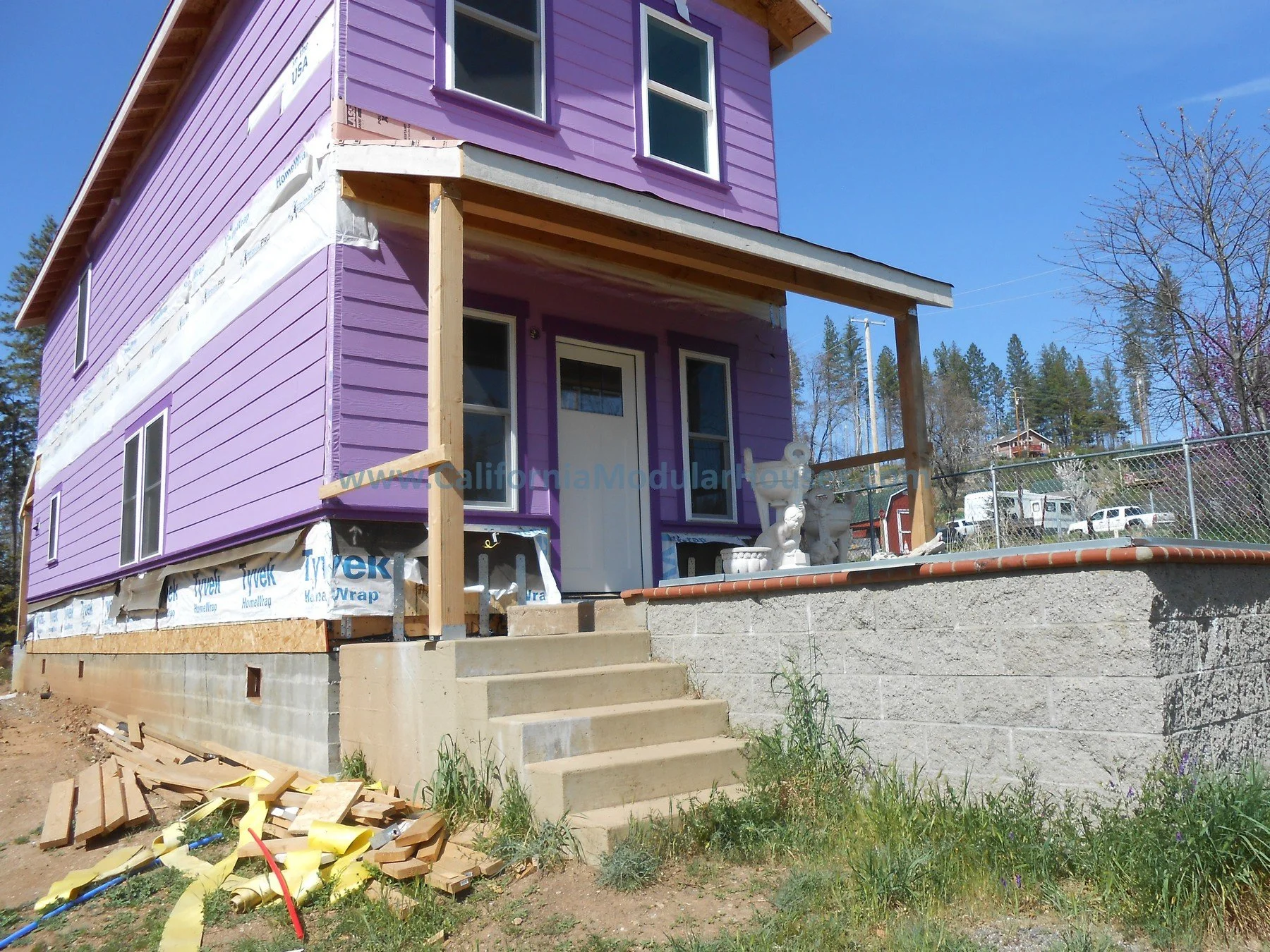 A two-story house under construction with purple siding and white window frames. It has a front porch with concrete stairs and decorative statues.  Cobb Mountain, Lake County, California.  Modular Home CA, Modular Homes, Pre-Fabricated Homes, Prefab 