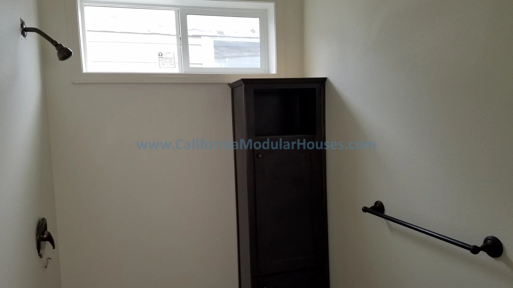 A small bathroom with a window at the top, a black wooden cabinet on the wall, and a black shower head and towel bar against the white walls.  California Modular, California Modular Home,  Modular Homes.