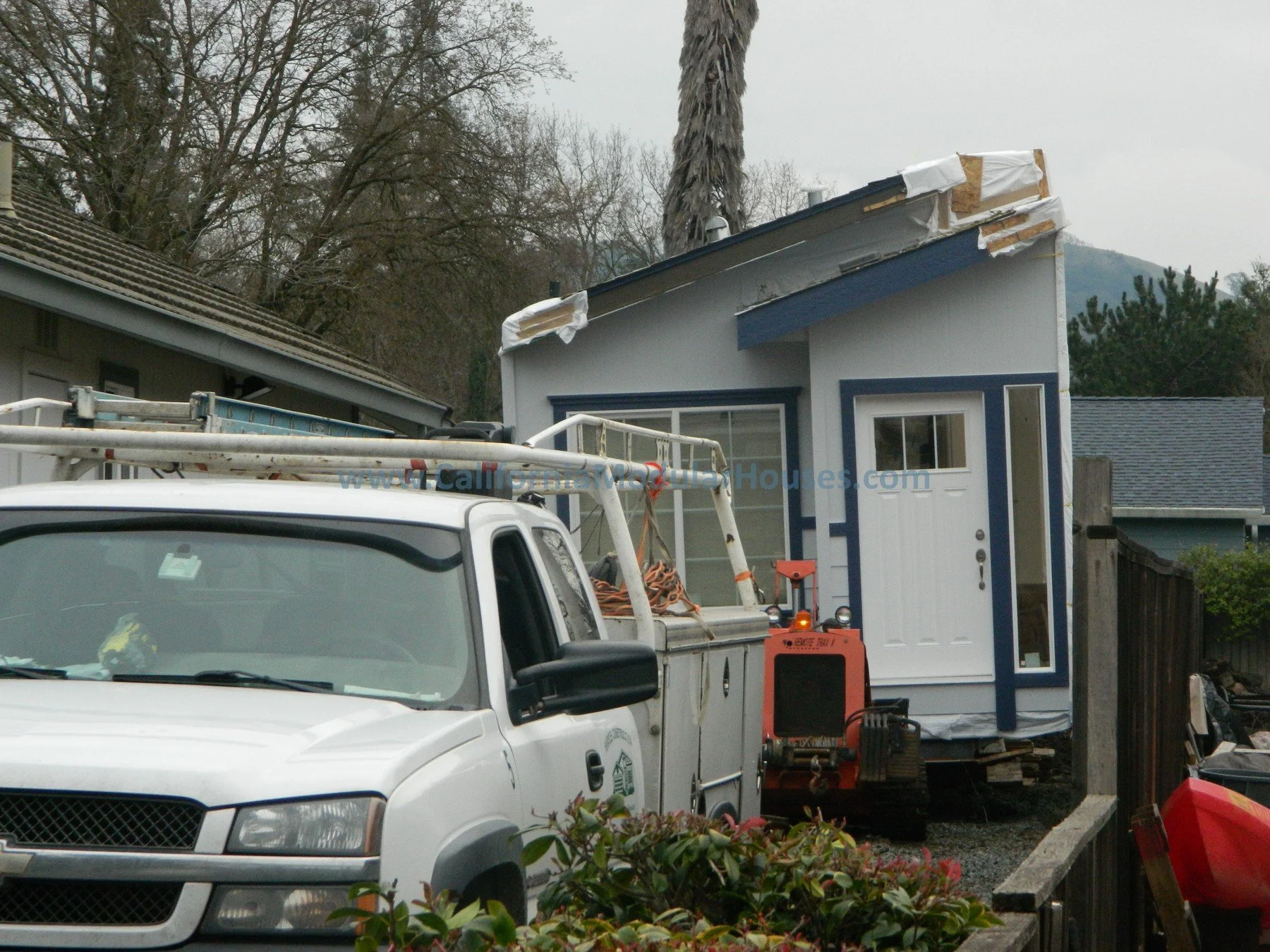 A white pickup truck with a bucket lift attached is parked in front of a small house that is under construction or renovation. The house has an ongoing roof repair with tarps and materials visible. There are trees in the background and other neighbor