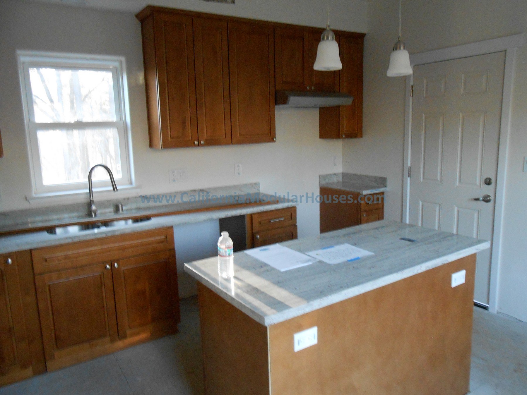 A kitchen with wooden cabinets, a window above the sink, a small kitchen island, pendant lights, and a door.  Cobb Mountain, Lake County, CA.  The San Ysidro Falls Two-Story model.  Modular Homes in California, Modular Home CA,