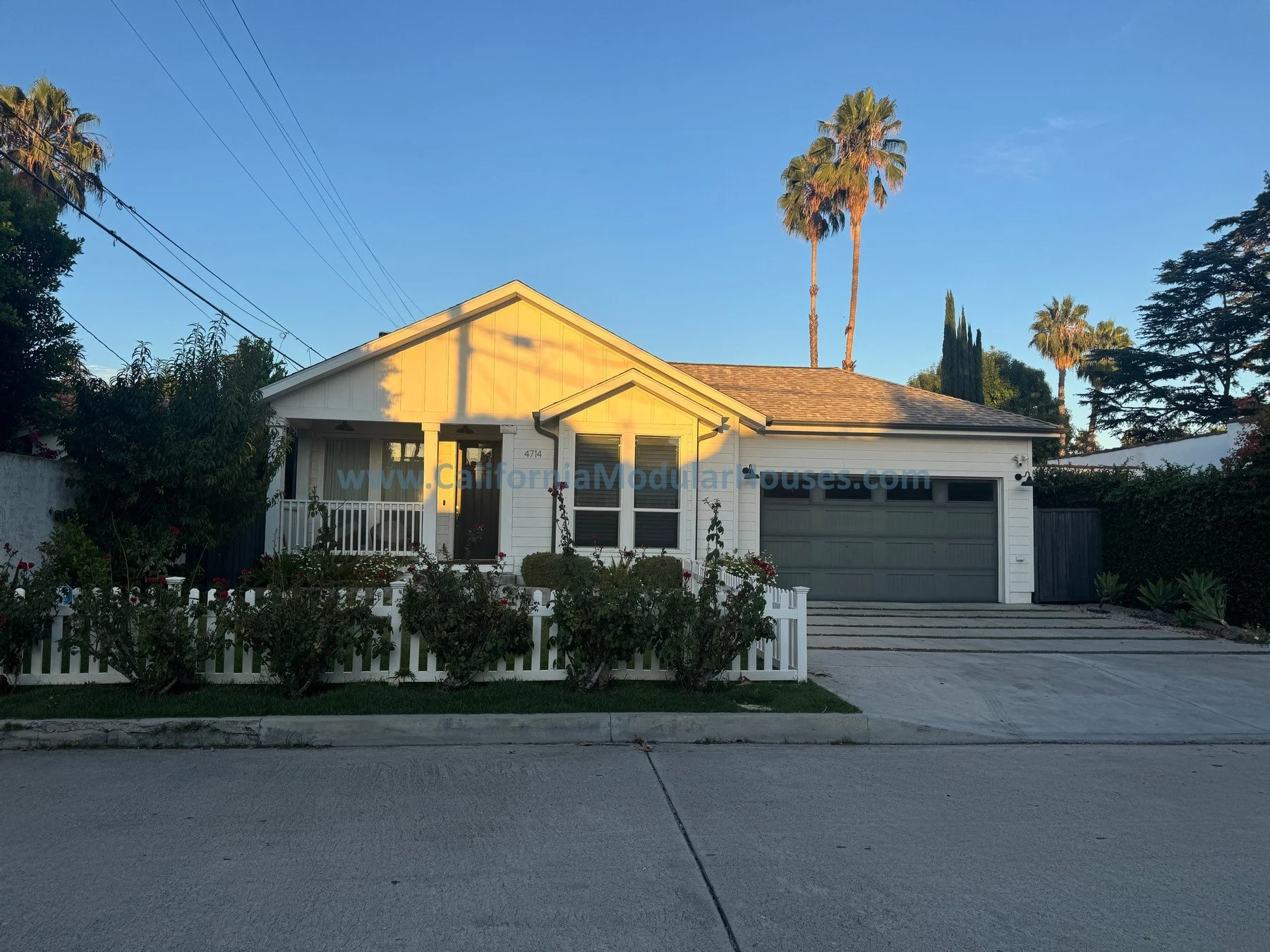 A white single-story house with a gray garage door, front porch, and a small white picket fence. The house has three large windows, a front door, and a gabled roof. There are flowering bushes in front of the fence and some tall palm trees and other t