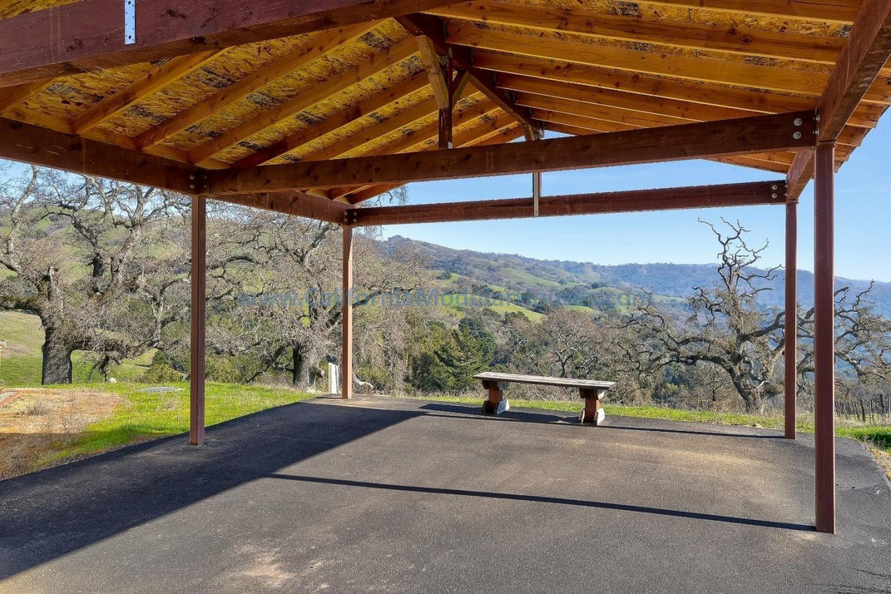 A covered outdoor pavilion with wooden beams and a bench, overlooking a scenic hilly landscape with leafless trees and distant hills under a clear blue sky. Bay Area Prefab Homes, Modular Homes.