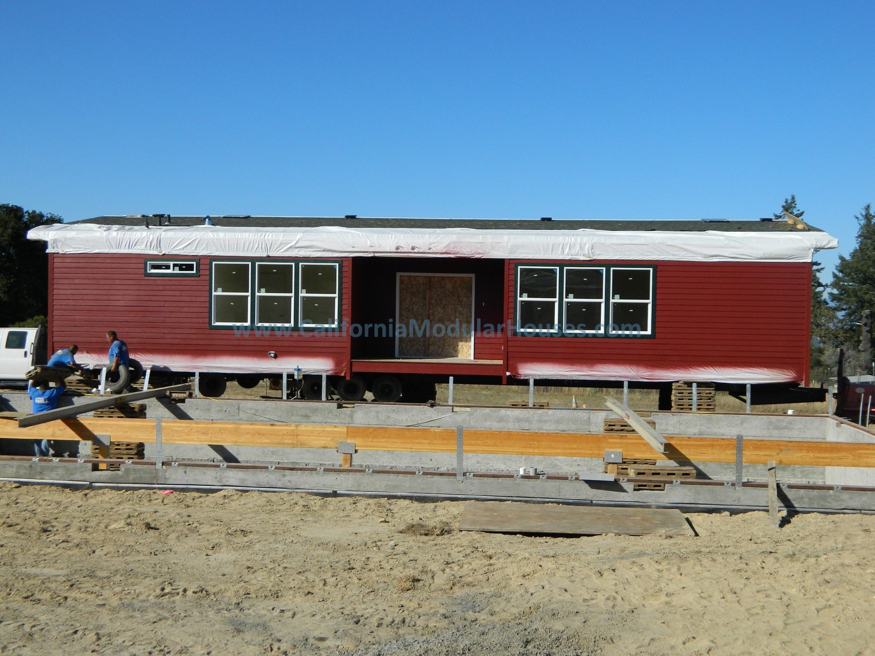 A red modular house under construction on a sandy lot, with workers assembling it and a clear blue sky overhead. California Prefab Modular Home, Modular Home CA.
