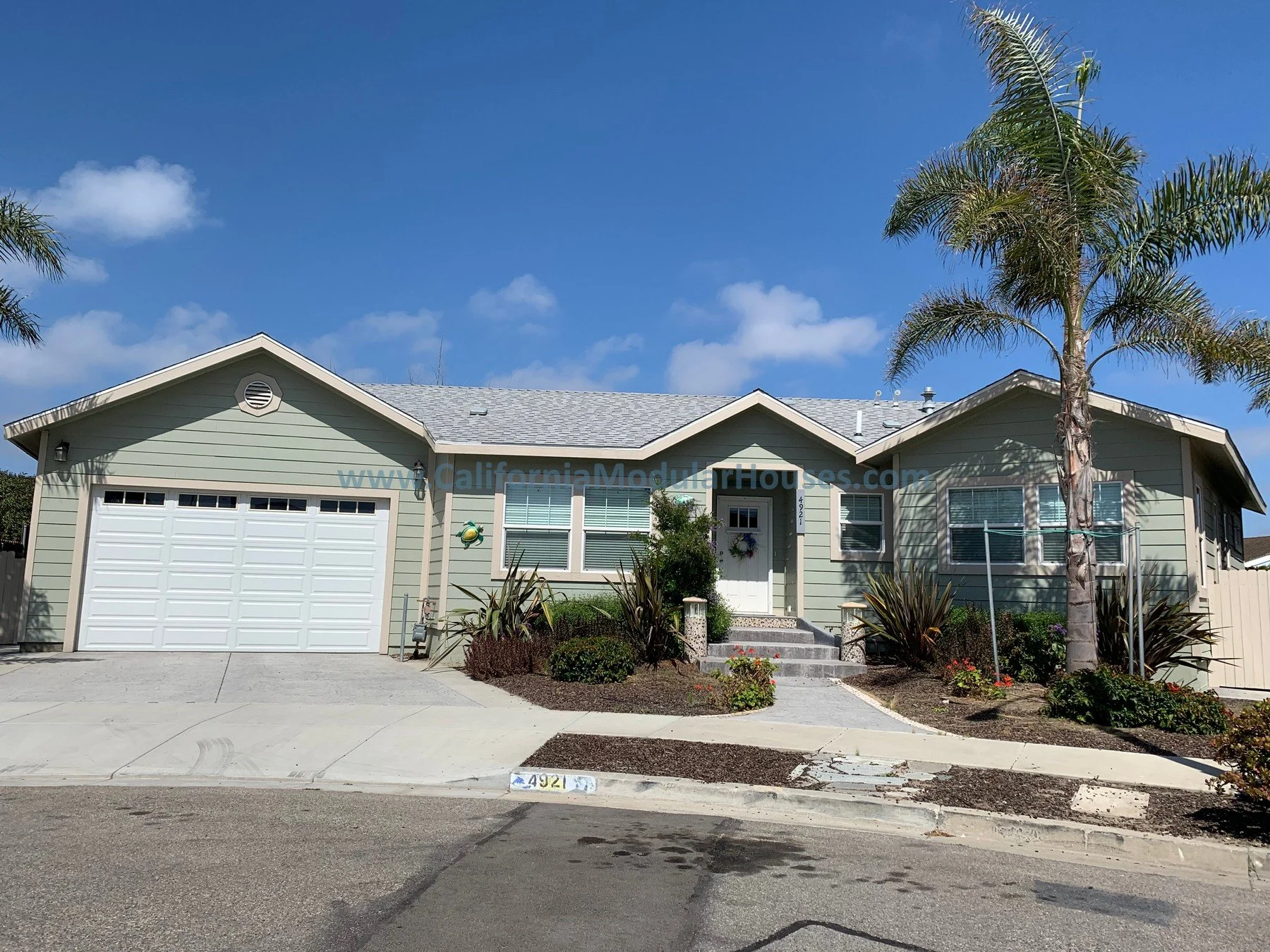 A single-story house with pale green siding, a gray shingle roof, a white garage door, and a small front yard with plants and bushes. Prefab modular home, Oxnard, Ventura County, CA.  California Modulars.  Modular Houses.  