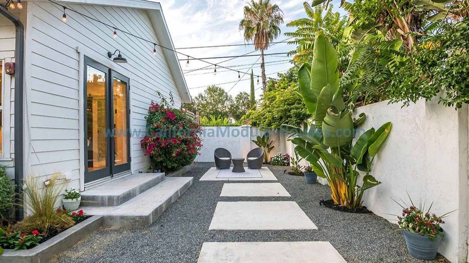 Outdoor patio area with white house siding, gray door with black trim, string lights above, and various green plants and flowers in pots along white fence wall. Centered paved pathway with stepping stones and seating area with two black chairs and sm