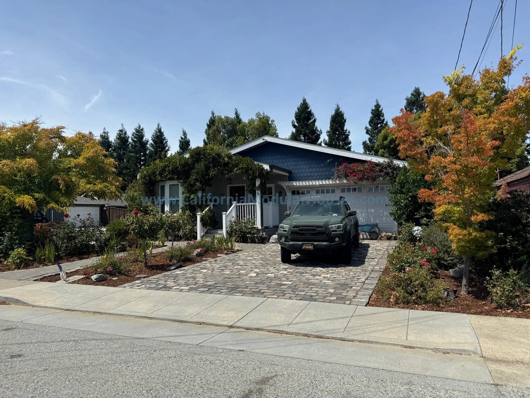 A suburban house with a driveway, surrounded by trees with autumn foliage, a black truck parked in the driveway, and a clear blue sky overhead.