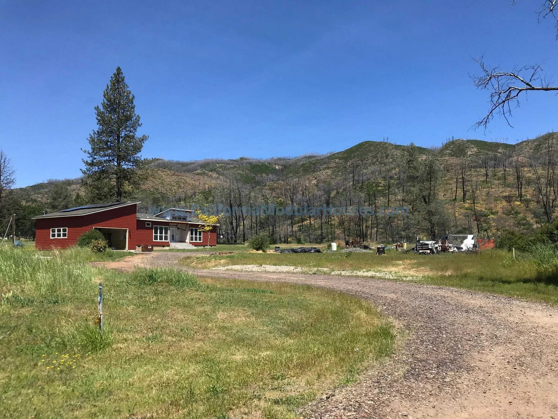 A red house with white trim sits on a grassy area with a gravel driveway in front, surrounded by green hills and trees under a clear blue sky.  Prefab Modular Homes California, California Modular.