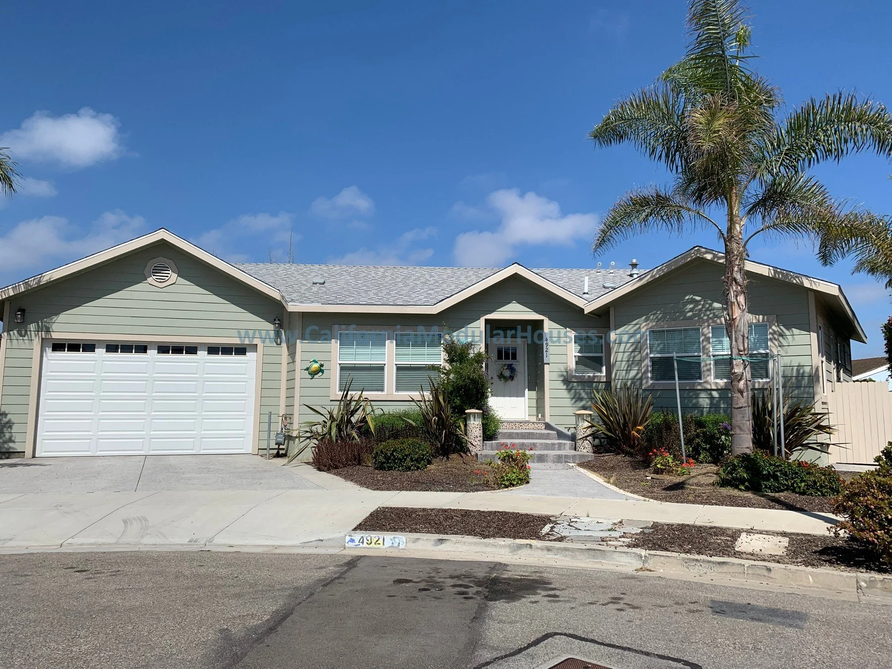 A light green, single-story house with a gray shingled roof, a two-car garage, a front porch with steps. California Modular Houses. Pre fab home.  Modular homes for California.  Casas prefabricadas.  Southern California.  