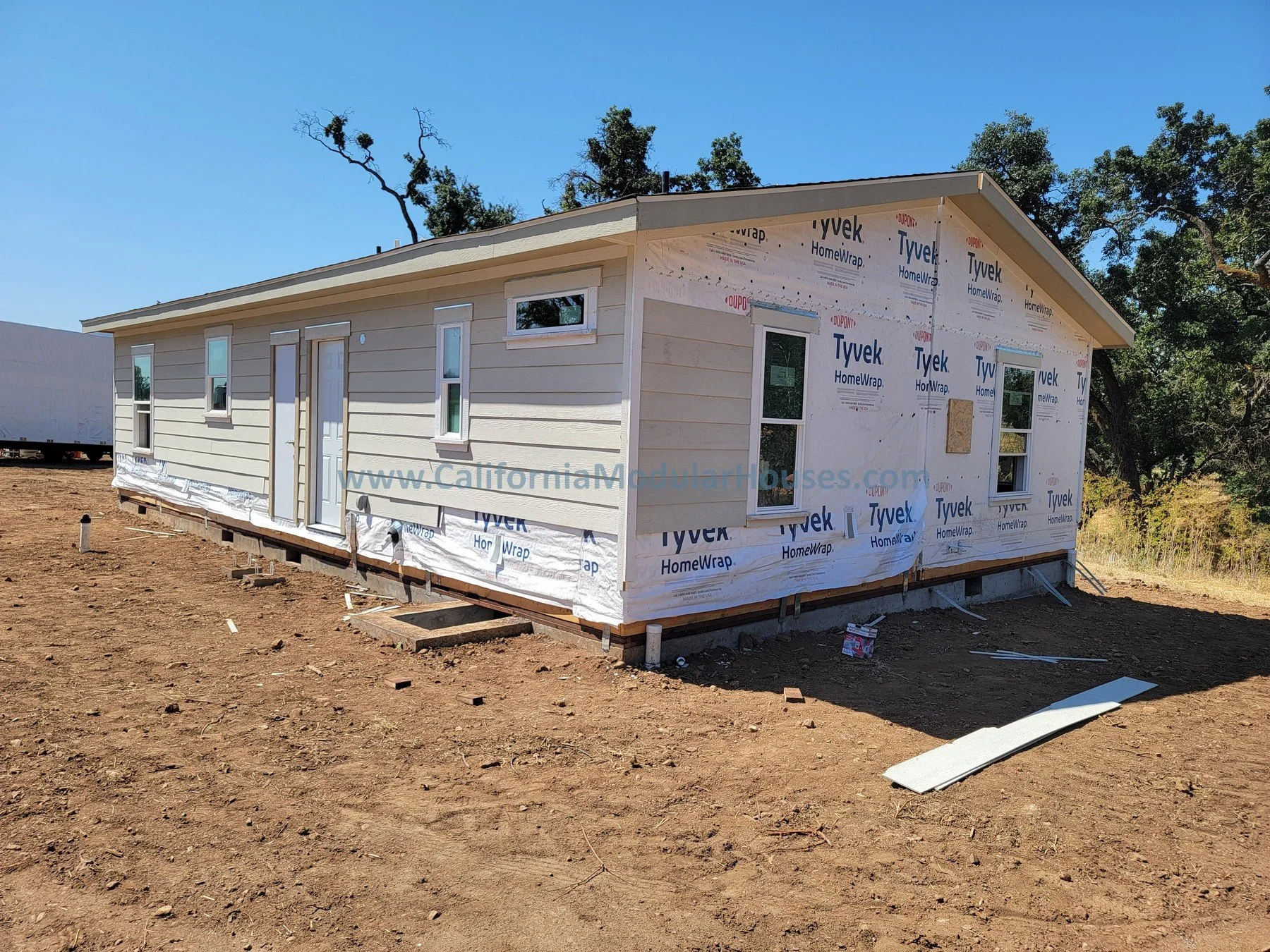 A house under construction with beige siding, multiple windows, and an unfinished exterior wall with Tyvek HomeWrap, on a dirt lot with a clear blue sky and trees in the background.