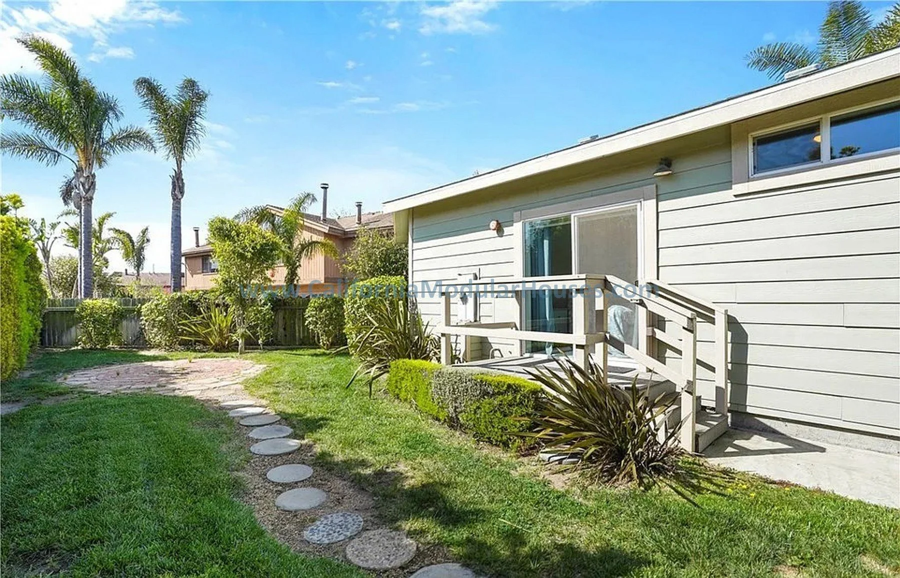 Backyard with a small wooden deck, sliding glass door, lush green grass, and various plants including palm trees.  California Modular Houses. Modular home construction California.  Prefab house.  Oxnard, Ventura County, CA.  