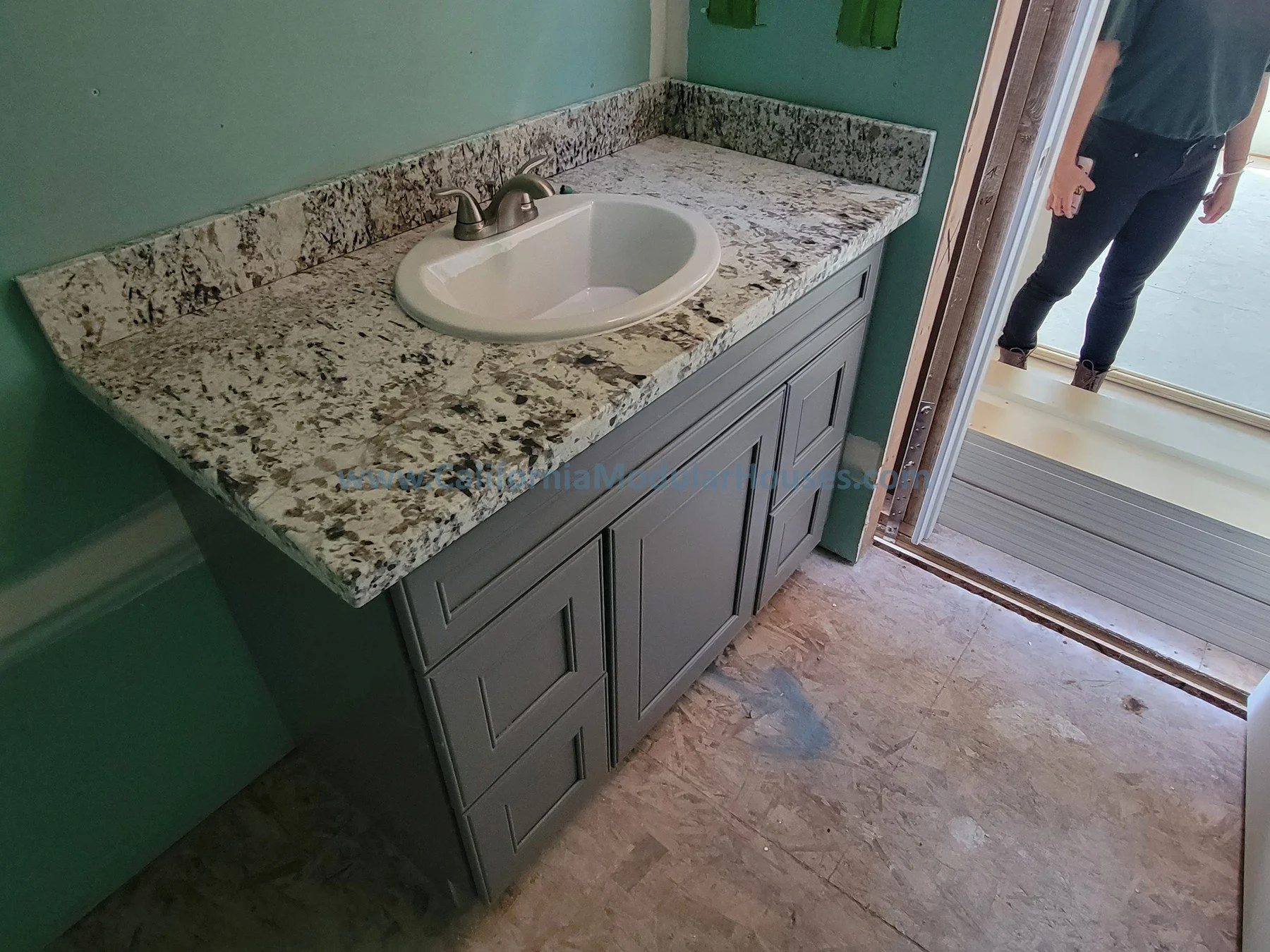 Bathroom vanity with granite countertop and a white oval sink, against green walls, partially open door showing a person standing outside.