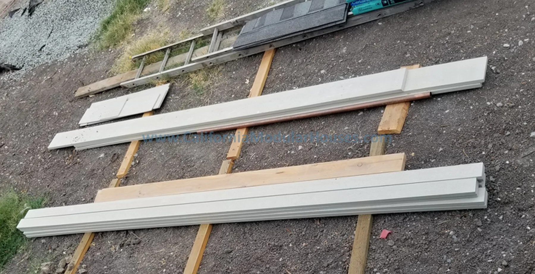 Construction materials including white siding, a ladder, and wooden beams laid on the ground outdoors.  California Modular Houses, Inc.  Modular granny flat.  California.   