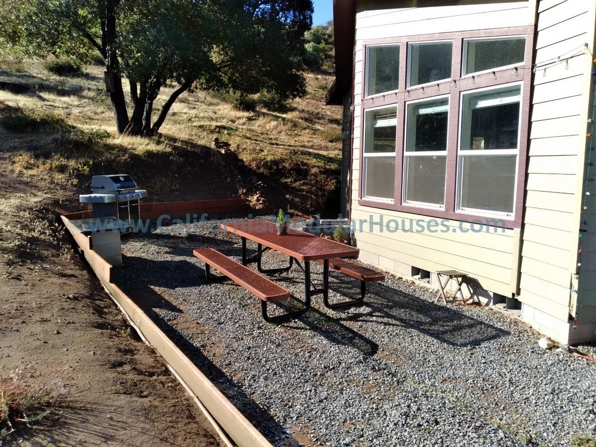 Backyard with a bbq grill, a red picnic table with benches, and a house with large window panes, surrounded by dirt and gravel ground.