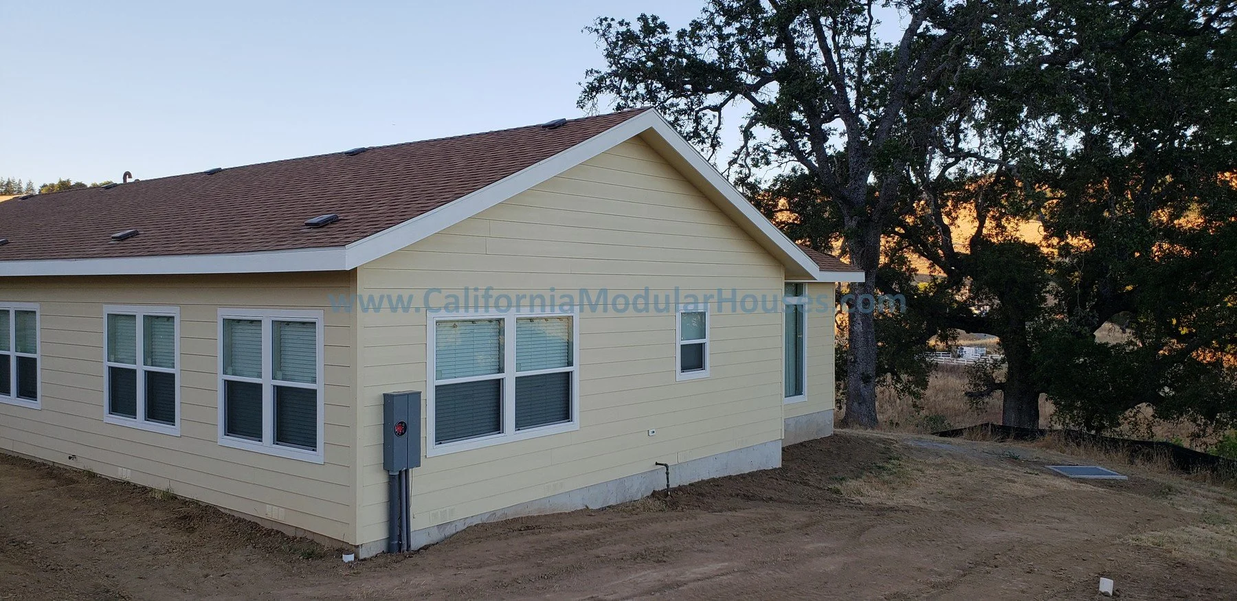 Side view of a newly built, yellow, single-story house with white trim, windows, and a brown shingle roof, set against a backdrop of trees at sunset. Bay Area Prefab Modular Home, Modular Home on a Sloped Property. 