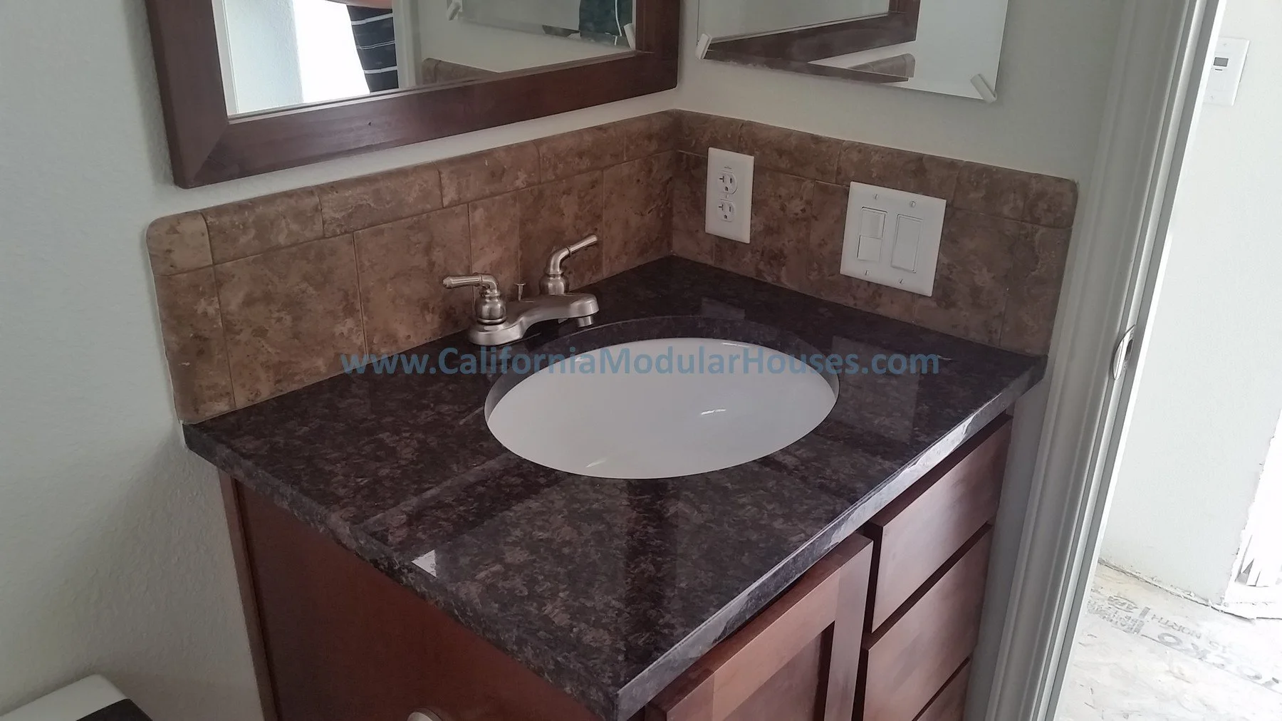 Bathroom vanity with a brown granite countertop, a white under-mount sink, two silver faucets, a brown tiled backsplash, a large mirror with a wooden frame, and electrical outlets on the wall.  Picture of Interior Modular Prefab ADU