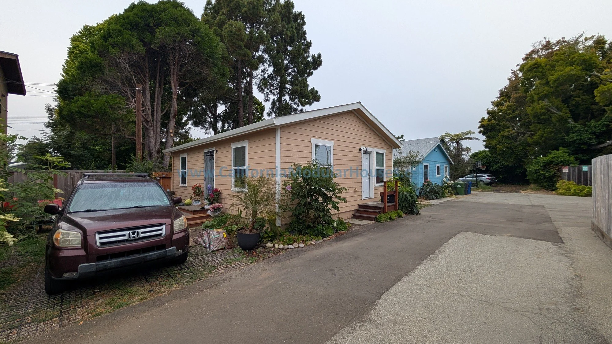 A residential area with a beige house with white trim, surrounded by plants and flowers, with a maroon SUV parked in front. There are tall trees behind the house and a blue house in the background, along with parked cars and a driveway.