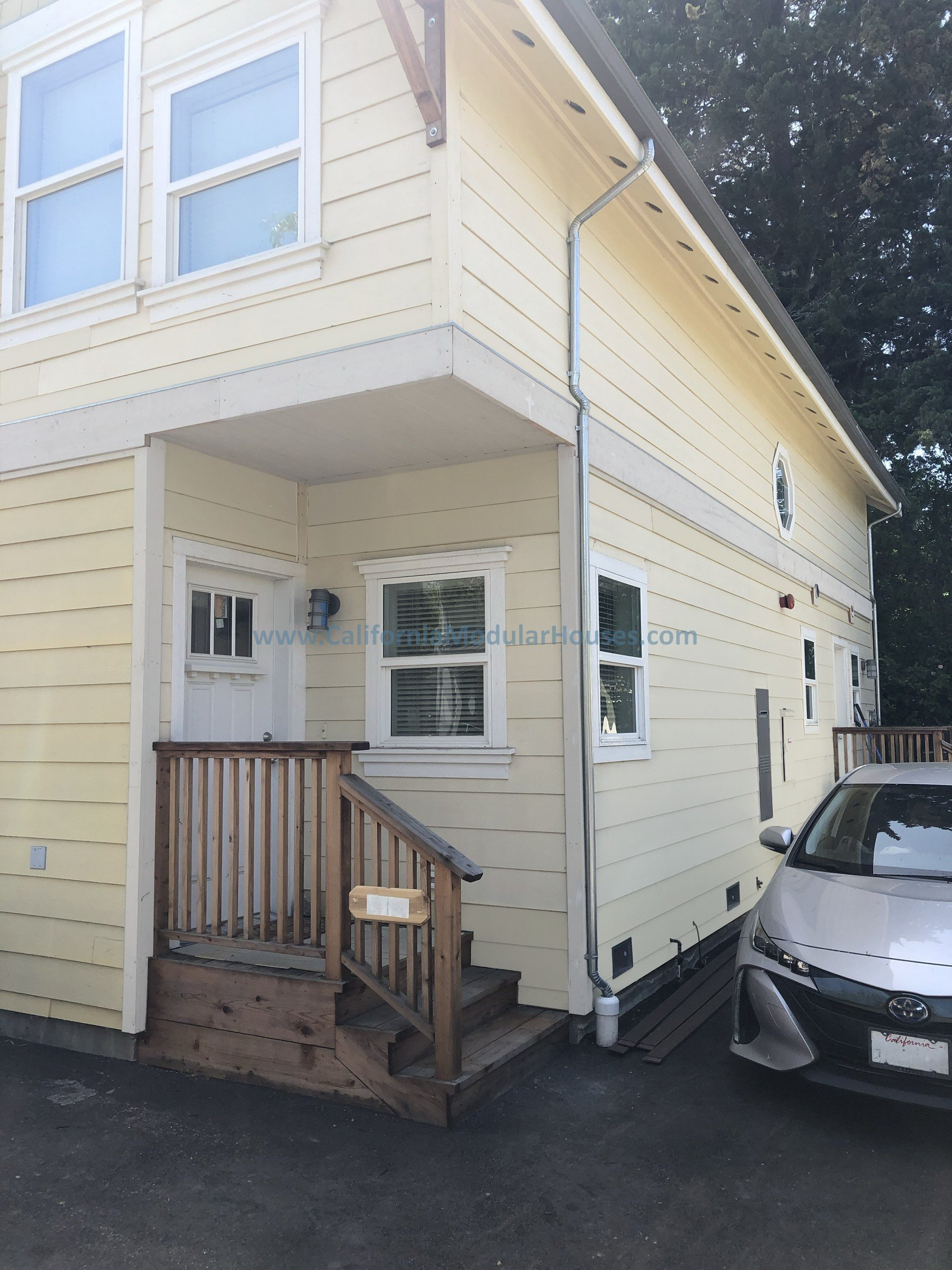 A two-story yellow house with white trim, a small wooden front porch with stairs, a gray car parked nearby, and a large tree in the background.