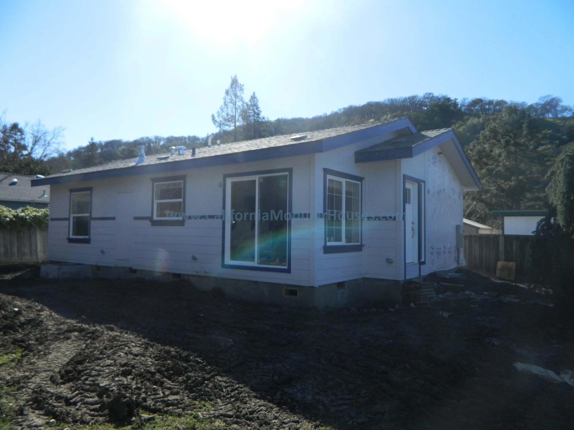 A house under construction with white siding, dark trim, and a sliding glass door, situated on a dirt foundation with a fenced backyard, set against a hillside with trees.