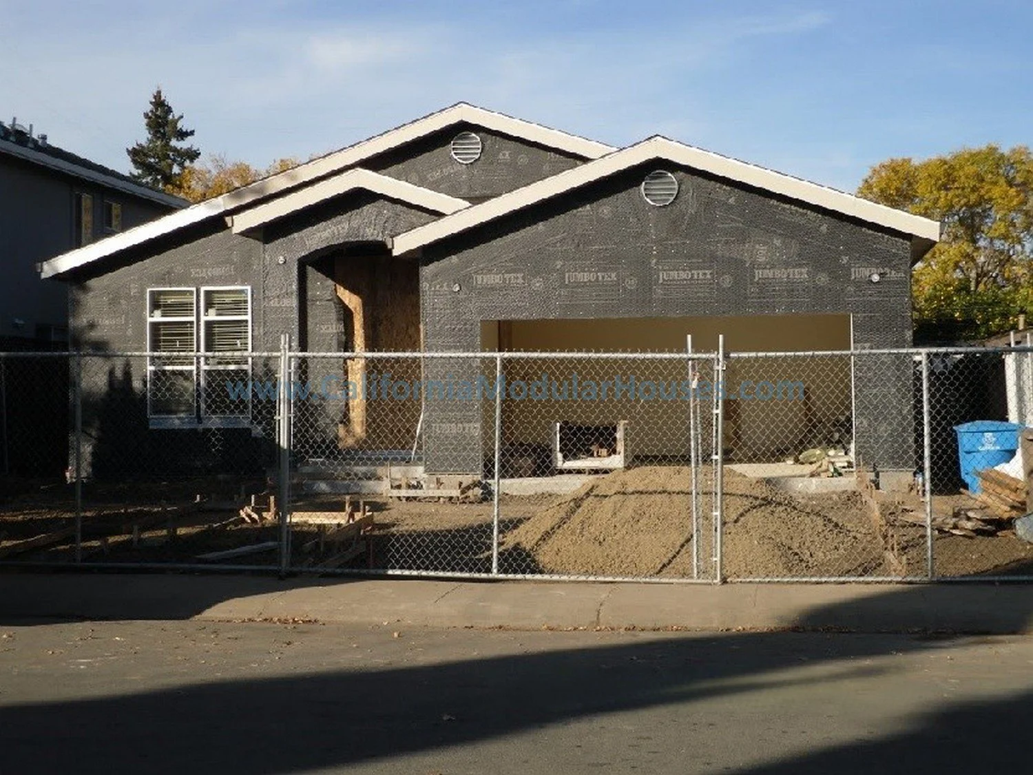 A house under construction with black roofing paper, boarded up windows, and a chain-link fence surrounding the property.