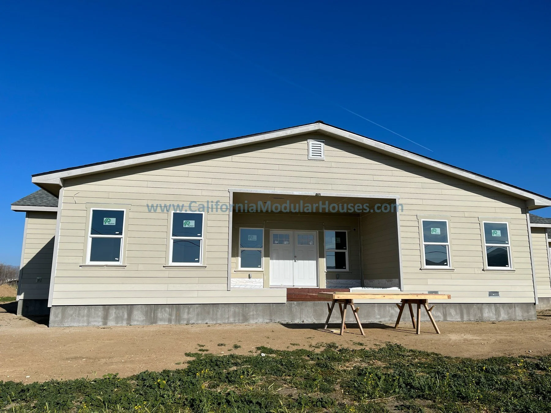 A house under construction with beige siding, four windows, a front door, and a partial porch, with a blue sky background.  California modular.  CA prefab.  