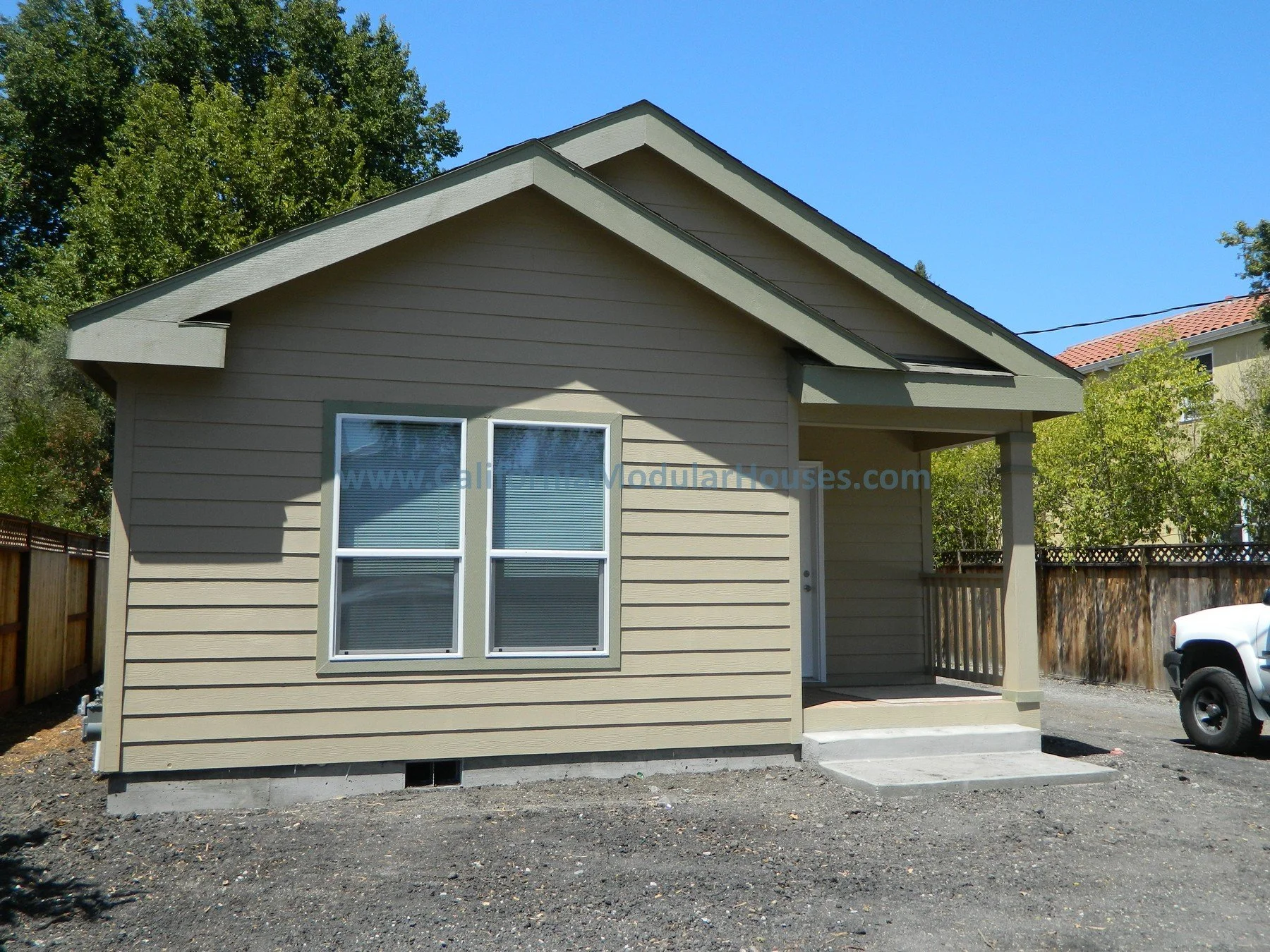 Front view of a small beige modular house with two windows, a small porch, and stairs, fenced yard, and a white truck parked nearby, under a clear blue sky.