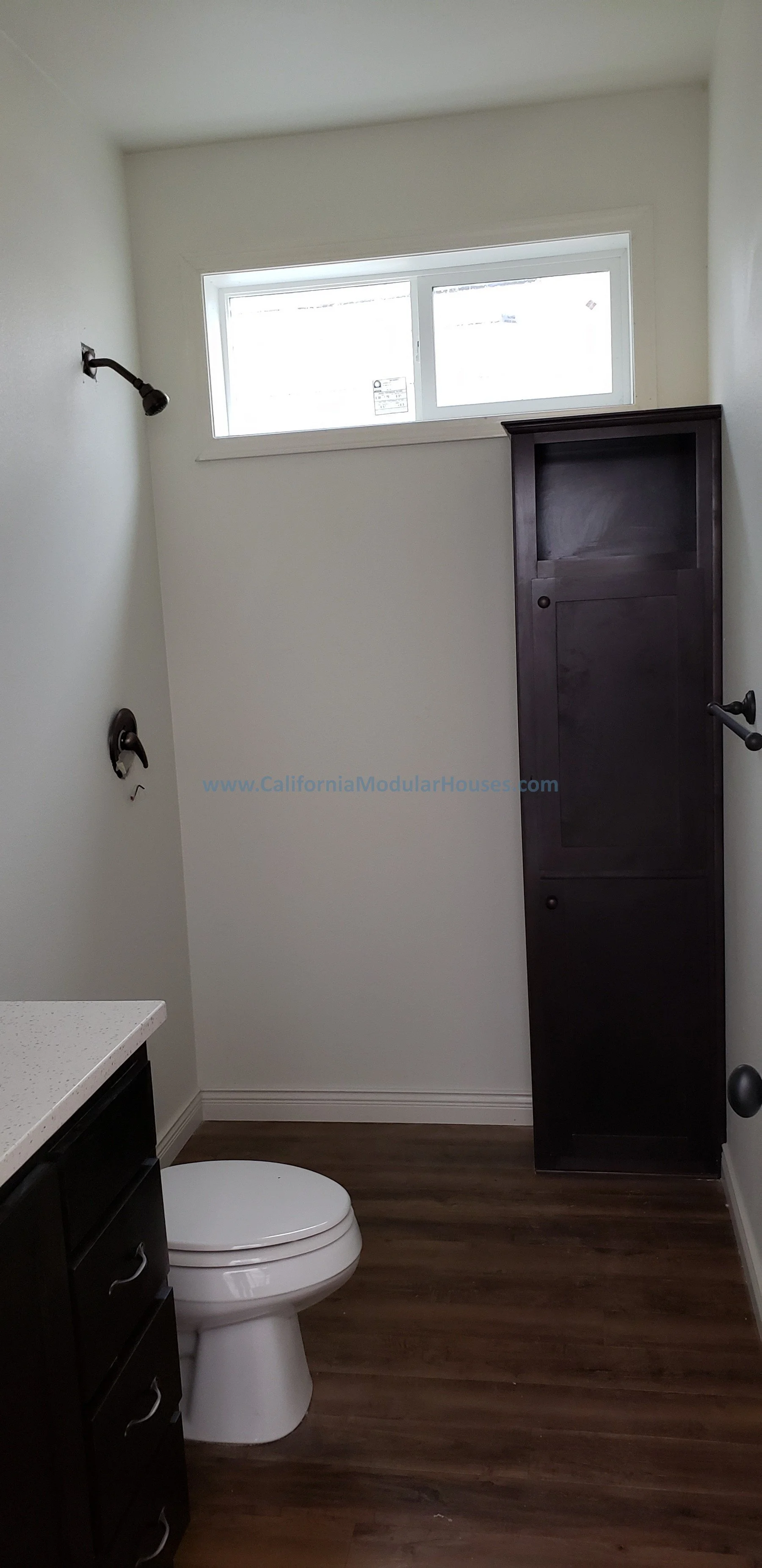 Small bathroom with a toilet, a dark wooden cabinet, a window near the ceiling, and a corner shower with no door.  Modular Home CA, California Modular Homes, Prefab Modular Homes California,