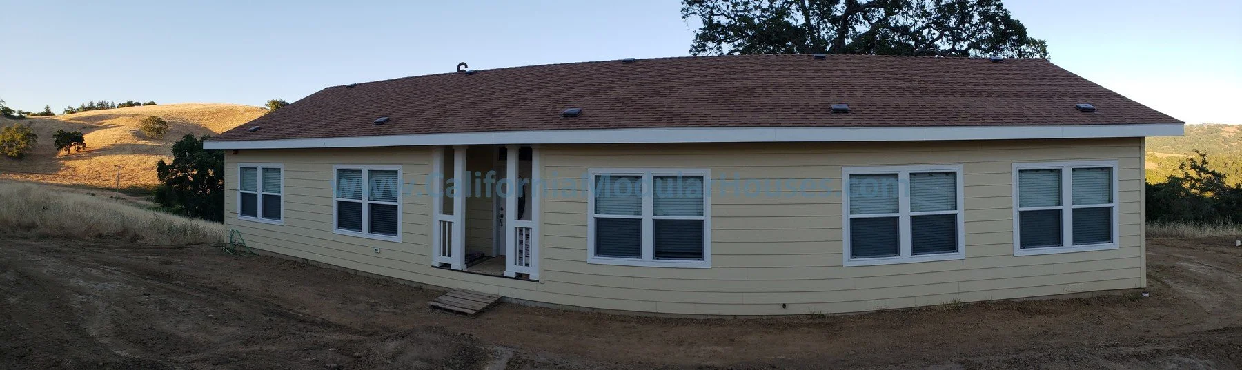 A single-story, beige house with white trim, located in a rural, hilly area, with multiple windows and a central entrance with a small porch and steps. California Modular Houses, Bay Area Prefab Modular Home, California Modular Homes,