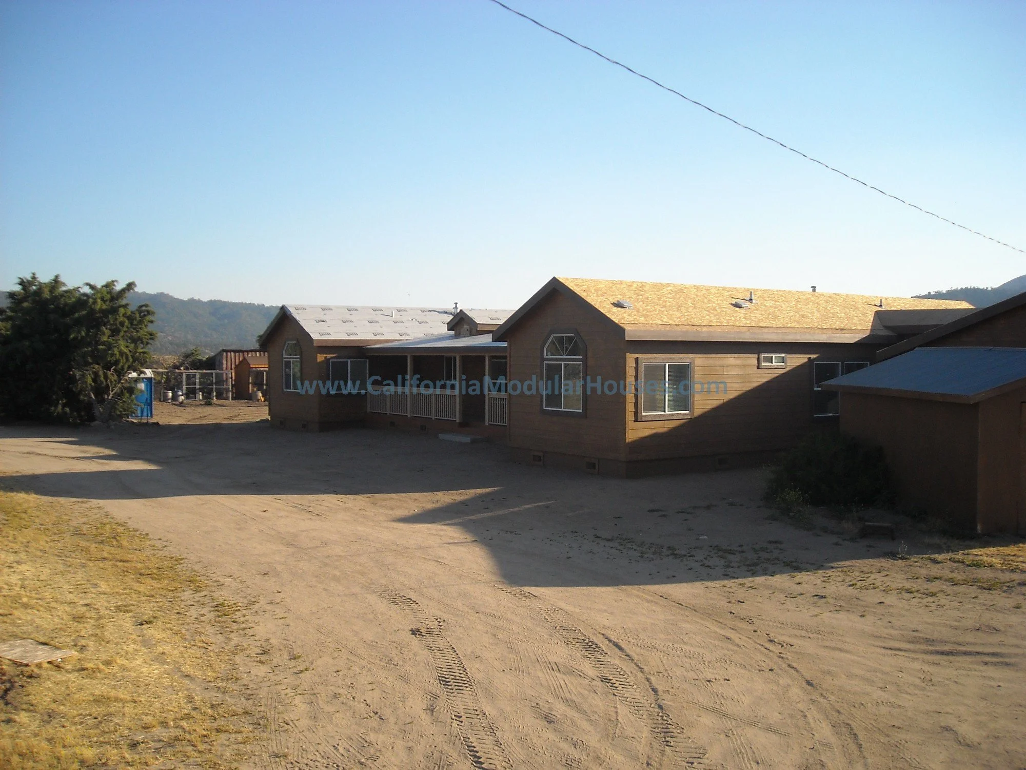 A brown modular house with a gabled roof in a rural area, dirt ground in the foreground, mountains in the background, and a blue sky overhead.