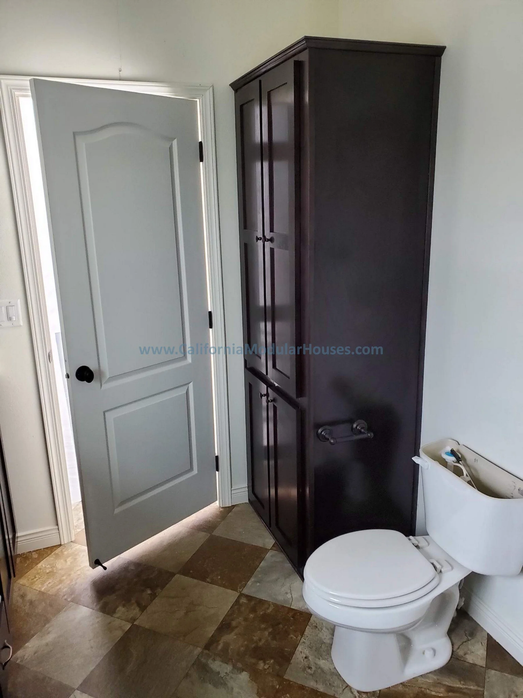 A bathroom with a partially open door, a white toilet with the tank lid removed, a dark tall cabinet next to the toilet, and a tiled floor with various shades of brown and beige.  California Modular, California Modular Home,  Modular Homes.