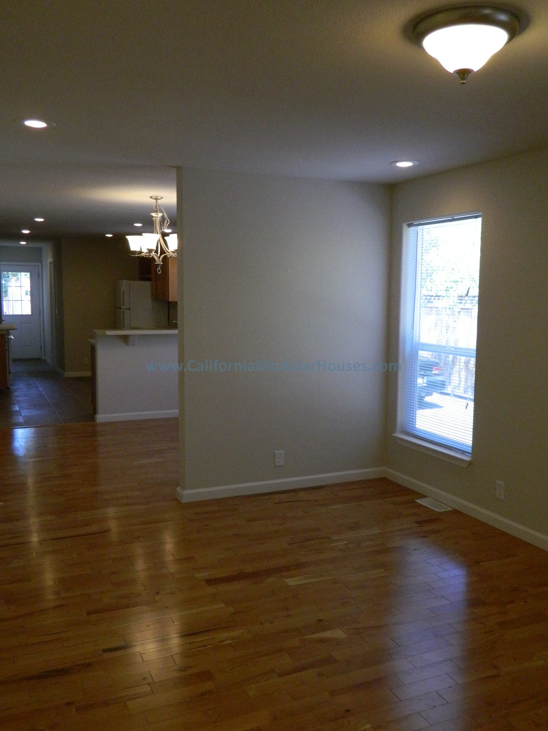 Empty living room with hardwood flooring, a large window, ceiling light, and an adjoining kitchen area with a chandelier and white appliances.
