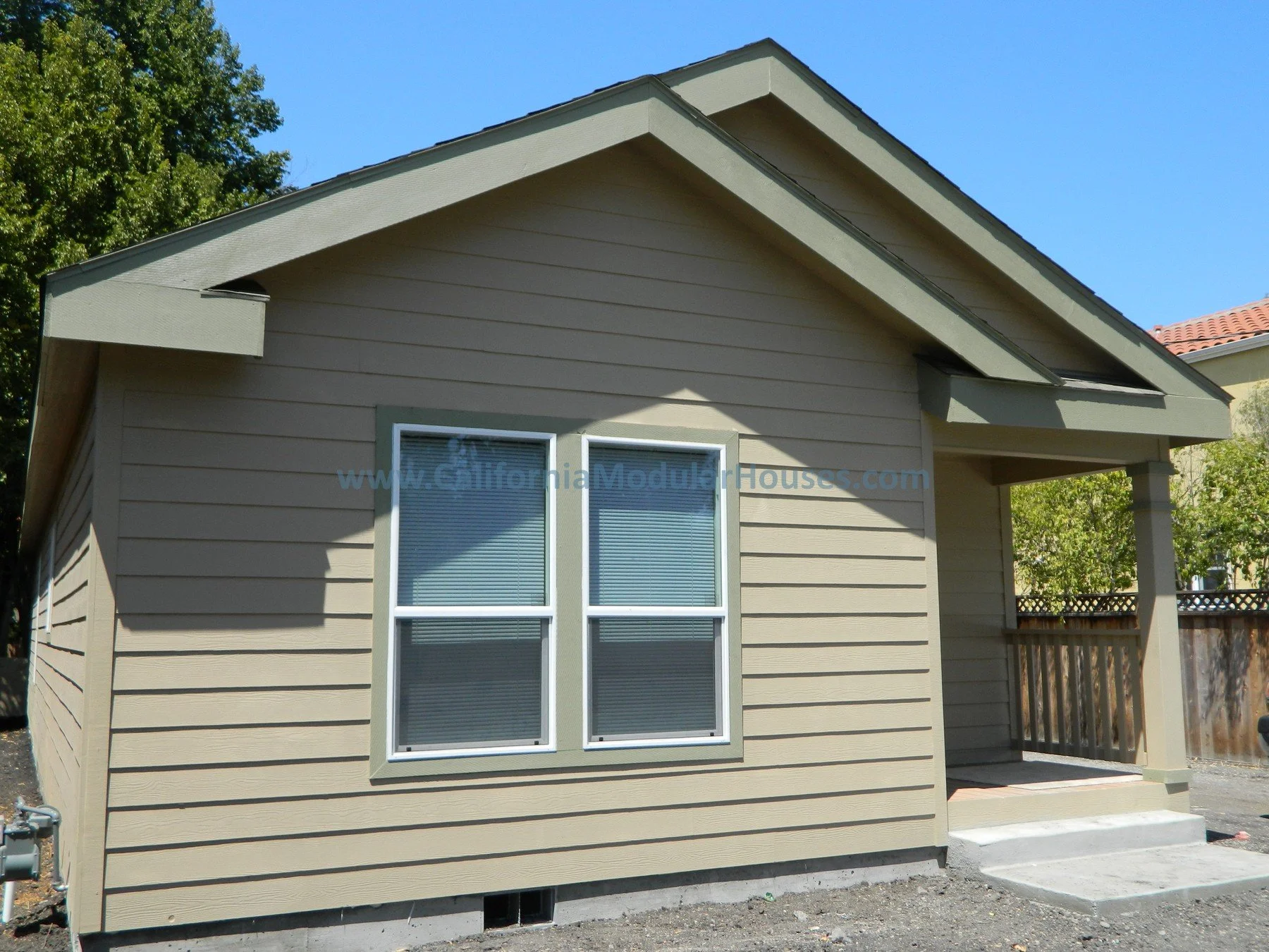 Front view of a small beige house with two windows and a porch with steps, surrounded by trees and a blue sky.
