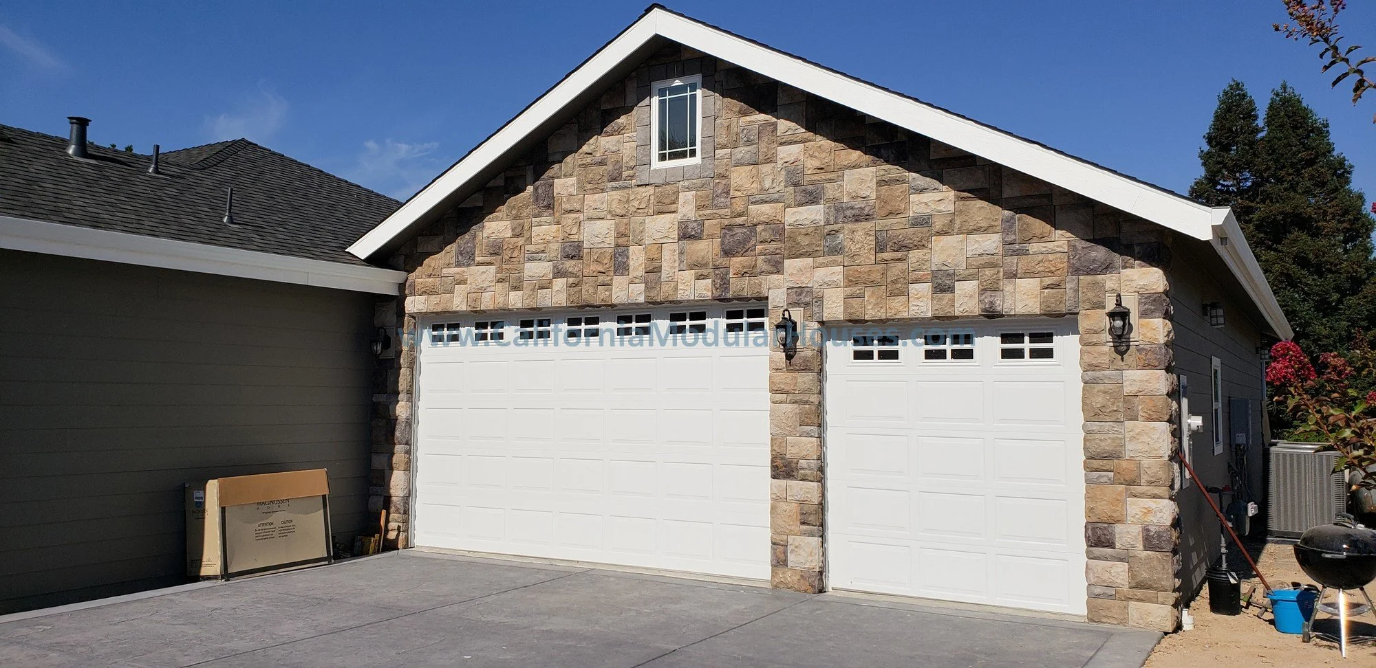 A detached garage with a stone facade and a white sectional garage door, situated at a residential property under a clear blue sky.  Factory Built Housing.  
