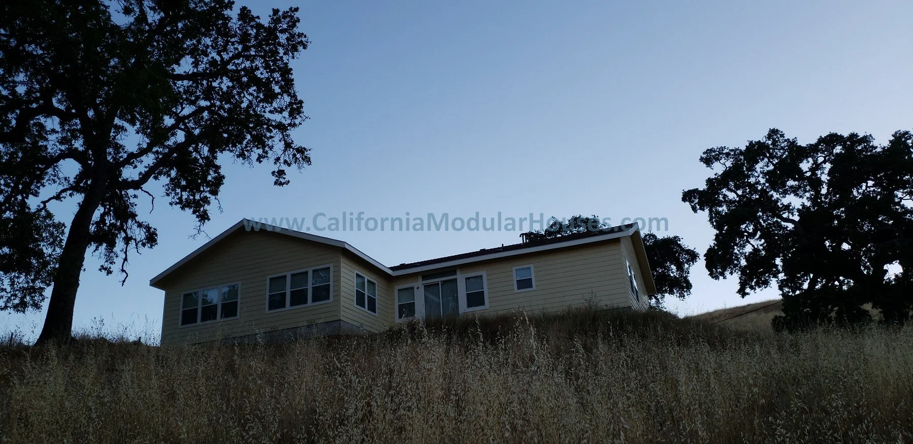 A beige house on a grassy hillside with trees on either side and a clear blue sky overhead. San Jose, Santa Clara County, CA. Modular Homes.