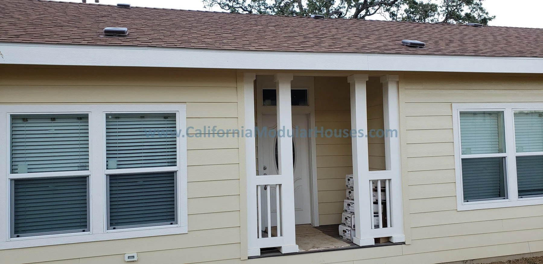 Exterior view of a yellow modular house under construction with two large windows with white frames and blinds, and an entrance with four white vertical posts and a door, with stacked boxes inside.  Modular Homes, Modular Home CA,
