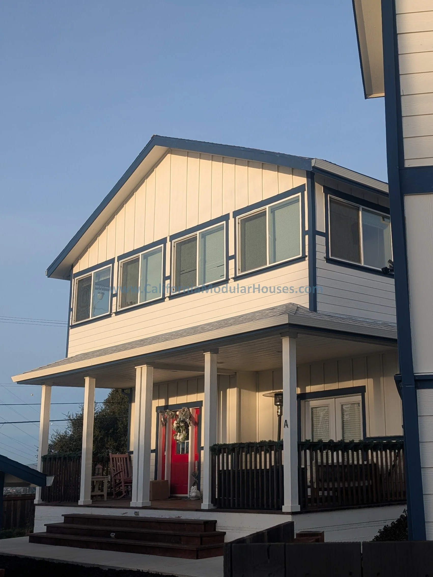A two-story modern modular house with a decorated front porch, a red door, and multiple windows, set against a clear sky.  Contra Costa County.  California East San Francisco Bay Modular Home.  Prefab Modular House.