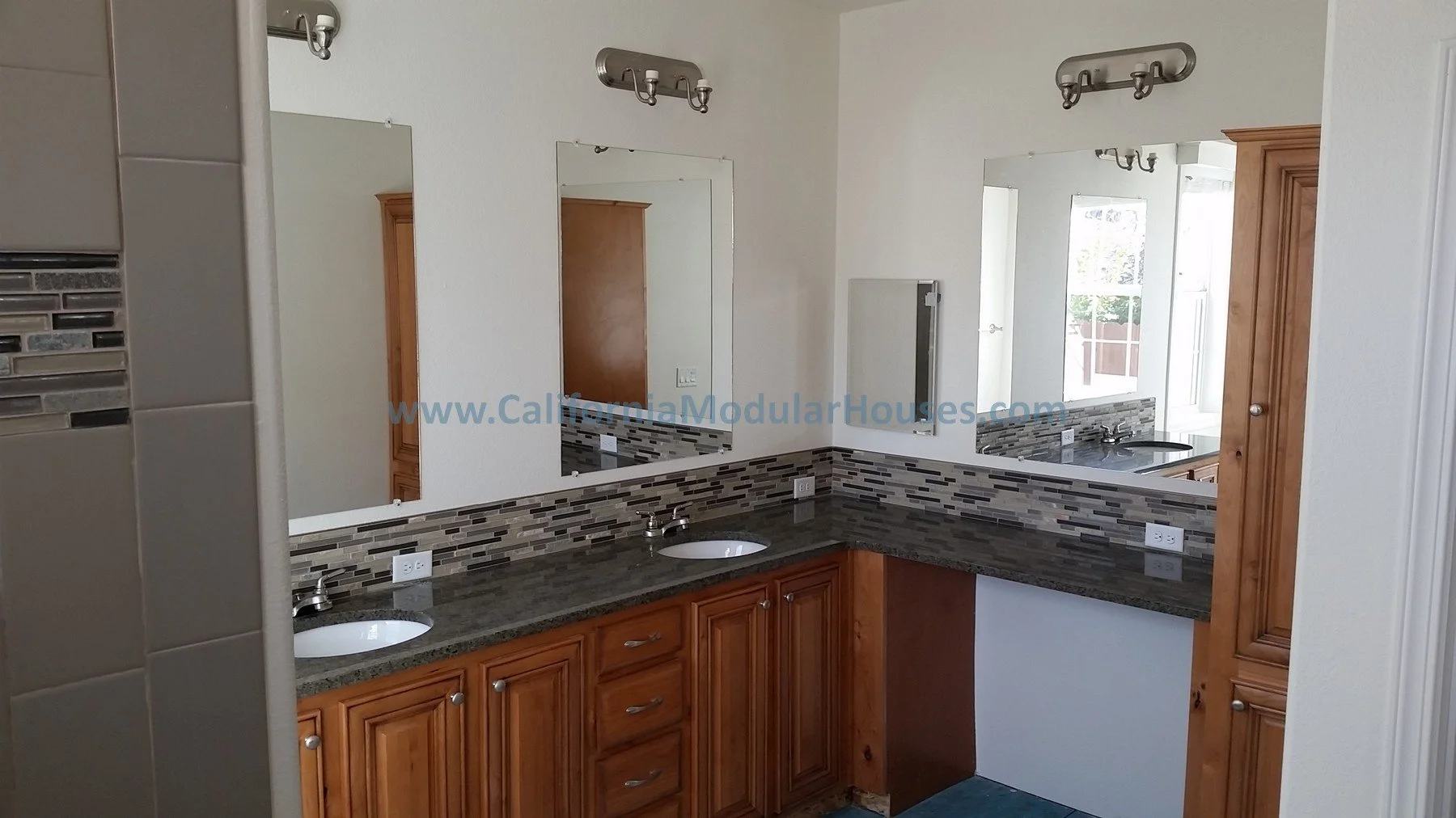 Bathroom with double vanity, granite countertops, wood cabinets, and mosaic tile backsplash, reflected in mirrors, with window and natural light.  Bay Area Prefab Homes, Modular Home CA.