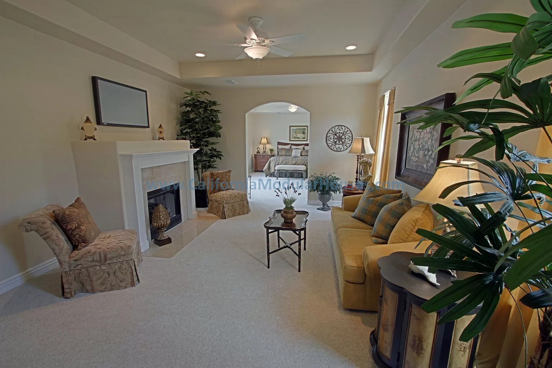 Living room with beige walls, yellow couch, patterned armchair, fireplace, framed art, ceiling fan, and potted plants.   Modular Homes CA. Prefab Homes California. Prefab Homes.