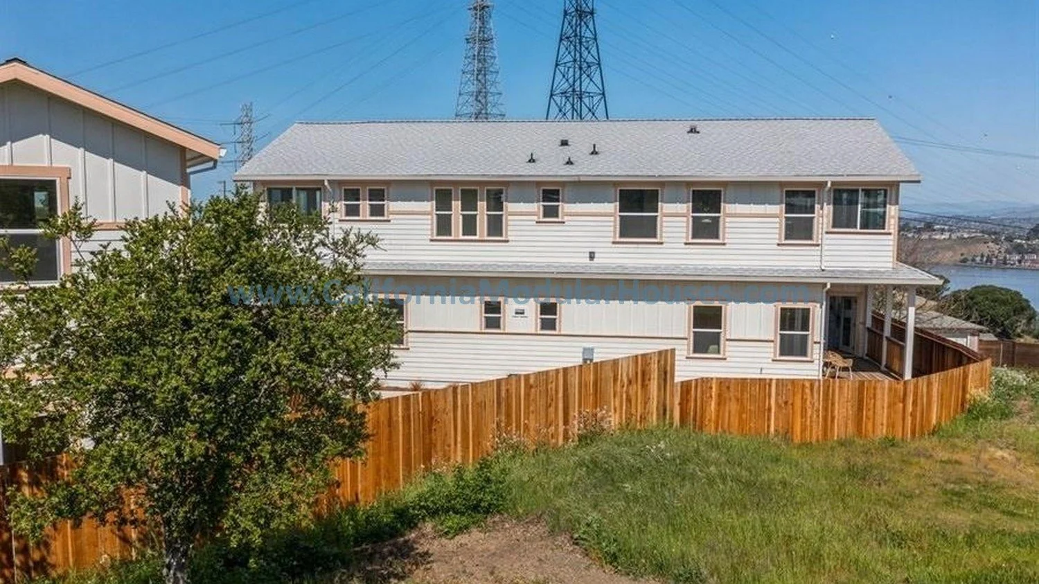 A white, two-story house with a gray roof, surrounded by a wooden fence, with a tree and a grassy yard in the foreground.  Two story modular single family modular residence.  Modular home.  