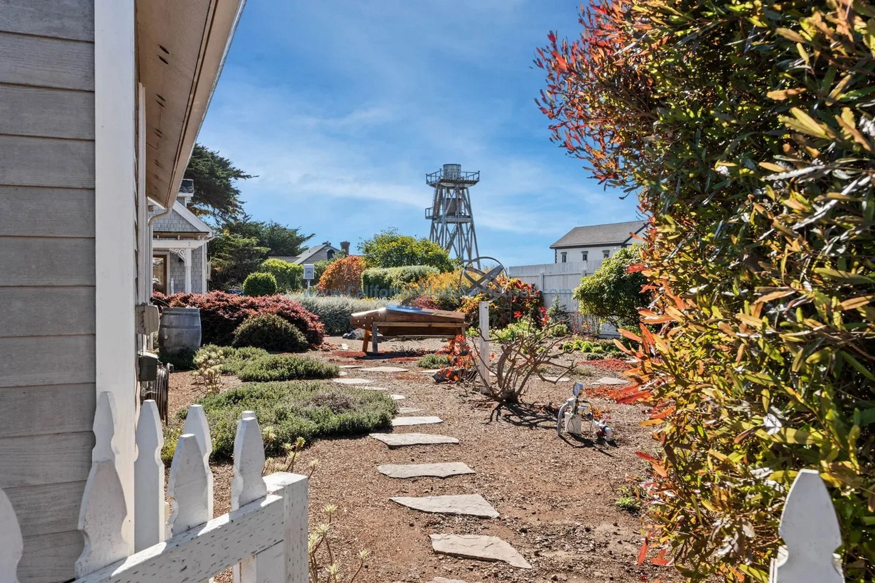 Mendocino, CA. Pre-fab modulars. Custom design coastal commission cape cod modular home. A garden with a stone path, a white picket fence, colorful bushes and plants, a bench, a small toy motorcycle, and a water tower.  