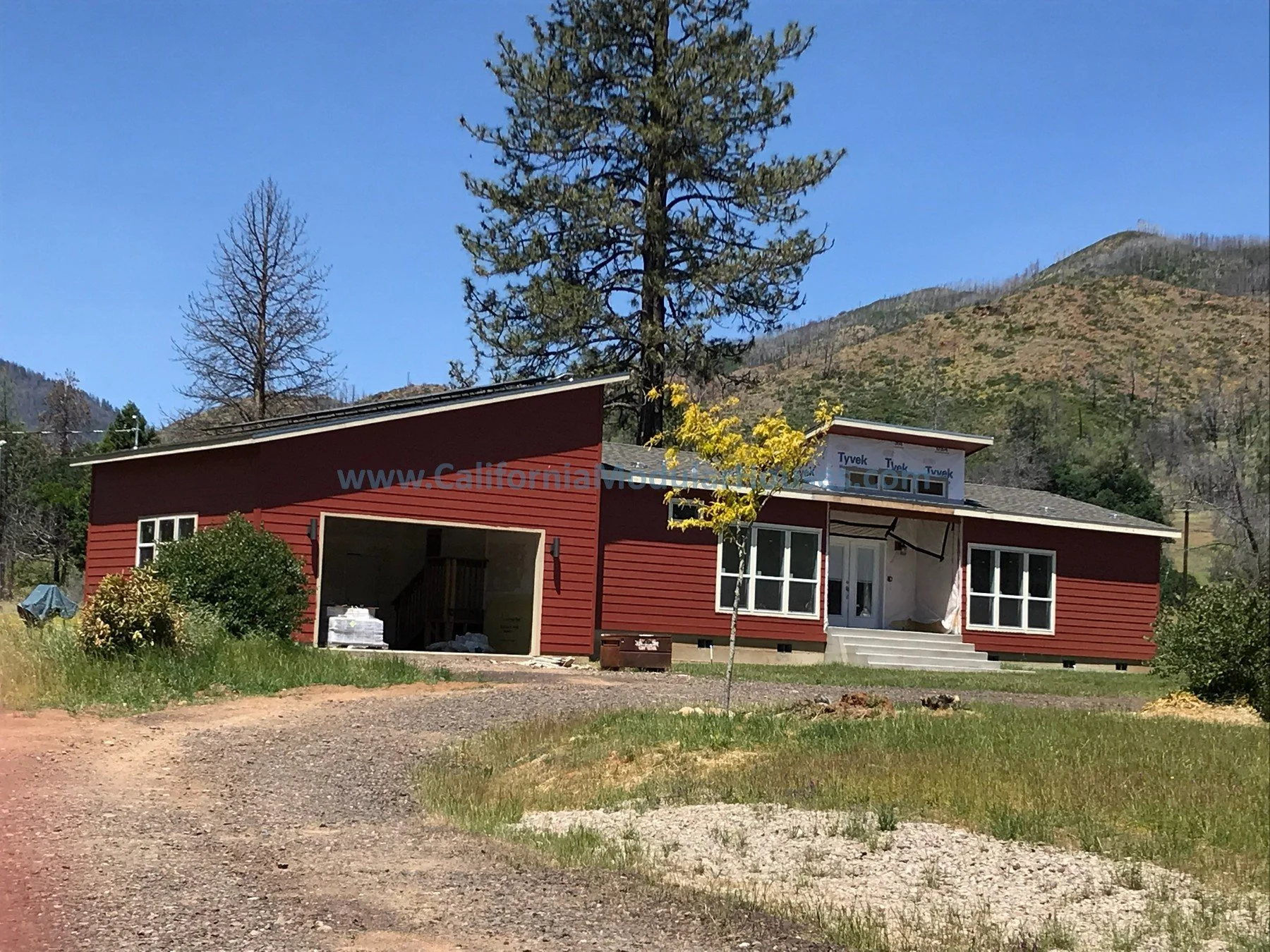A house under renovation with a red exterior, surrounded by trees and a dirt driveway, set against a mountainous landscape on a clear day.  Middletown, Lake County, CA. Lake County Modular.